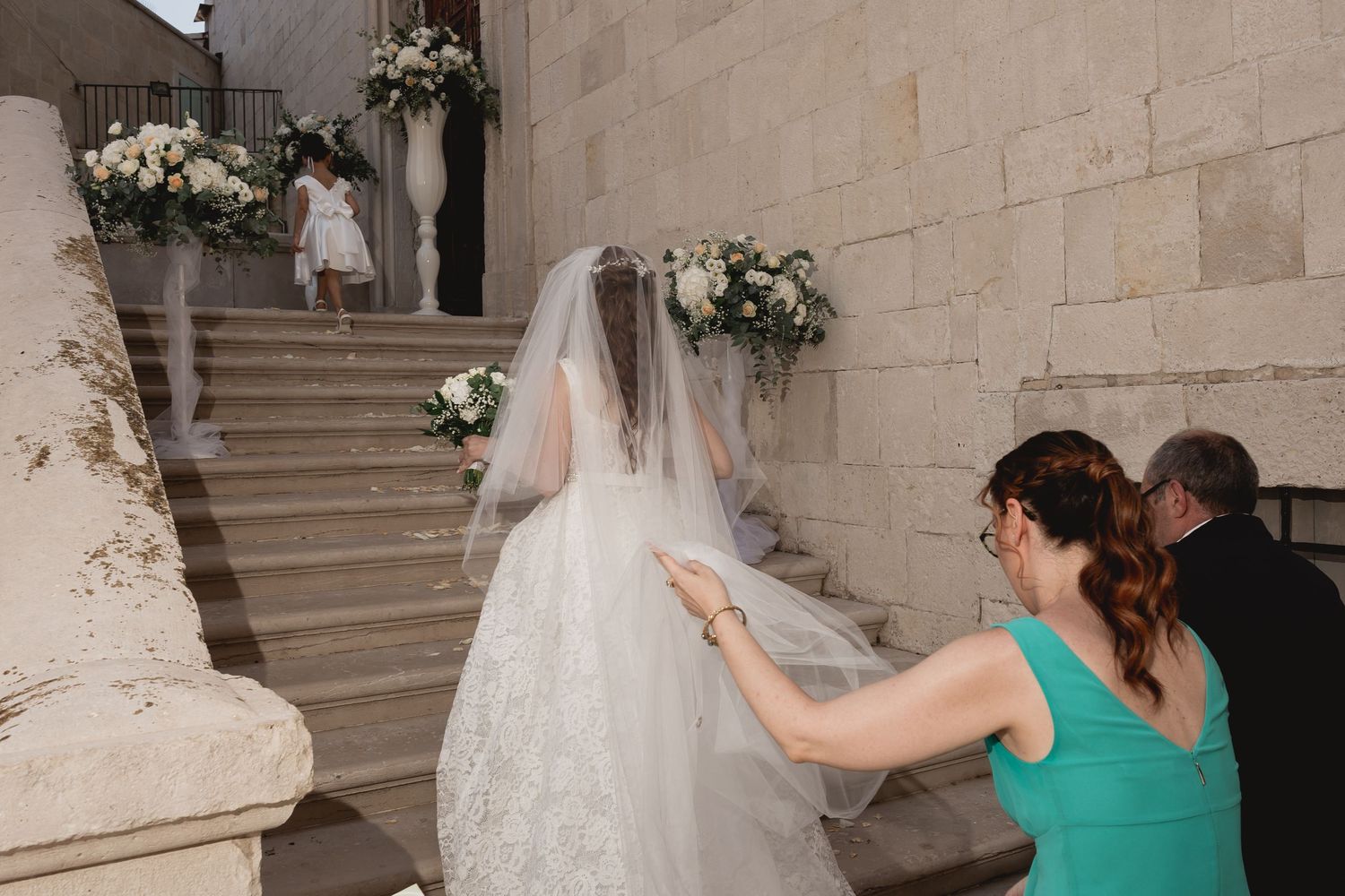 Wedding dress train and veil being adjusted on stone staircase with floral decorations.