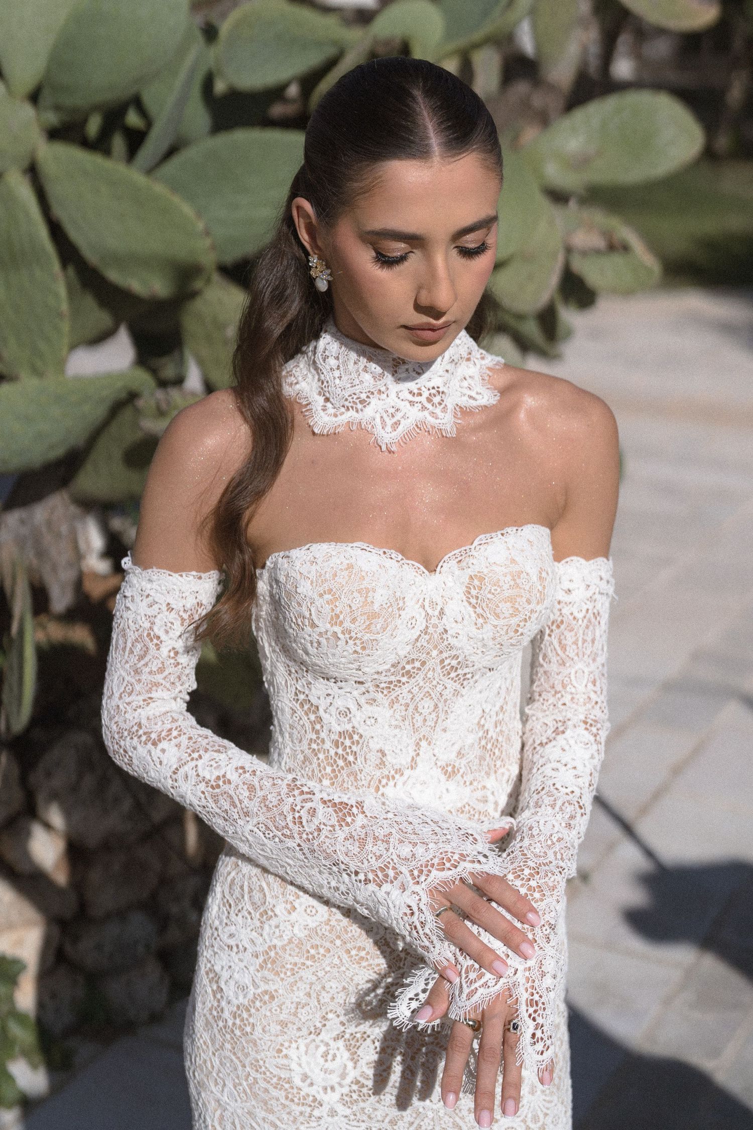 Bride in an off-shoulder lace wedding gown with sheer sleeves and a lace choker collar poses beside prickly pear cacti.