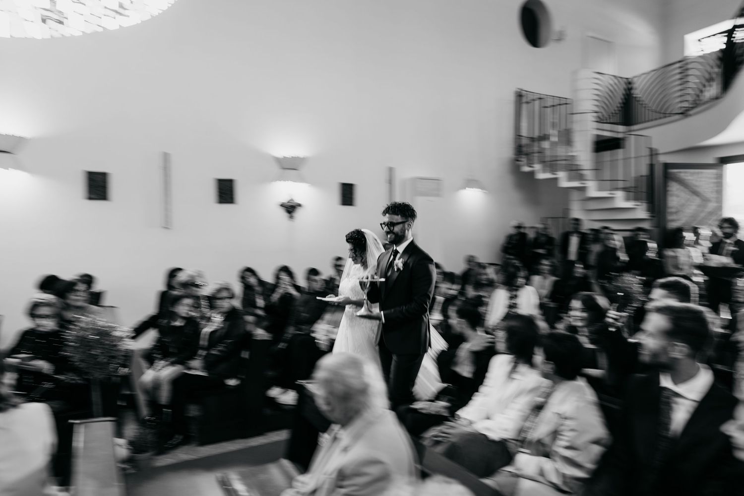 Bride and groom walk down the aisle past seated guests during an indoor wedding ceremony in black and white.