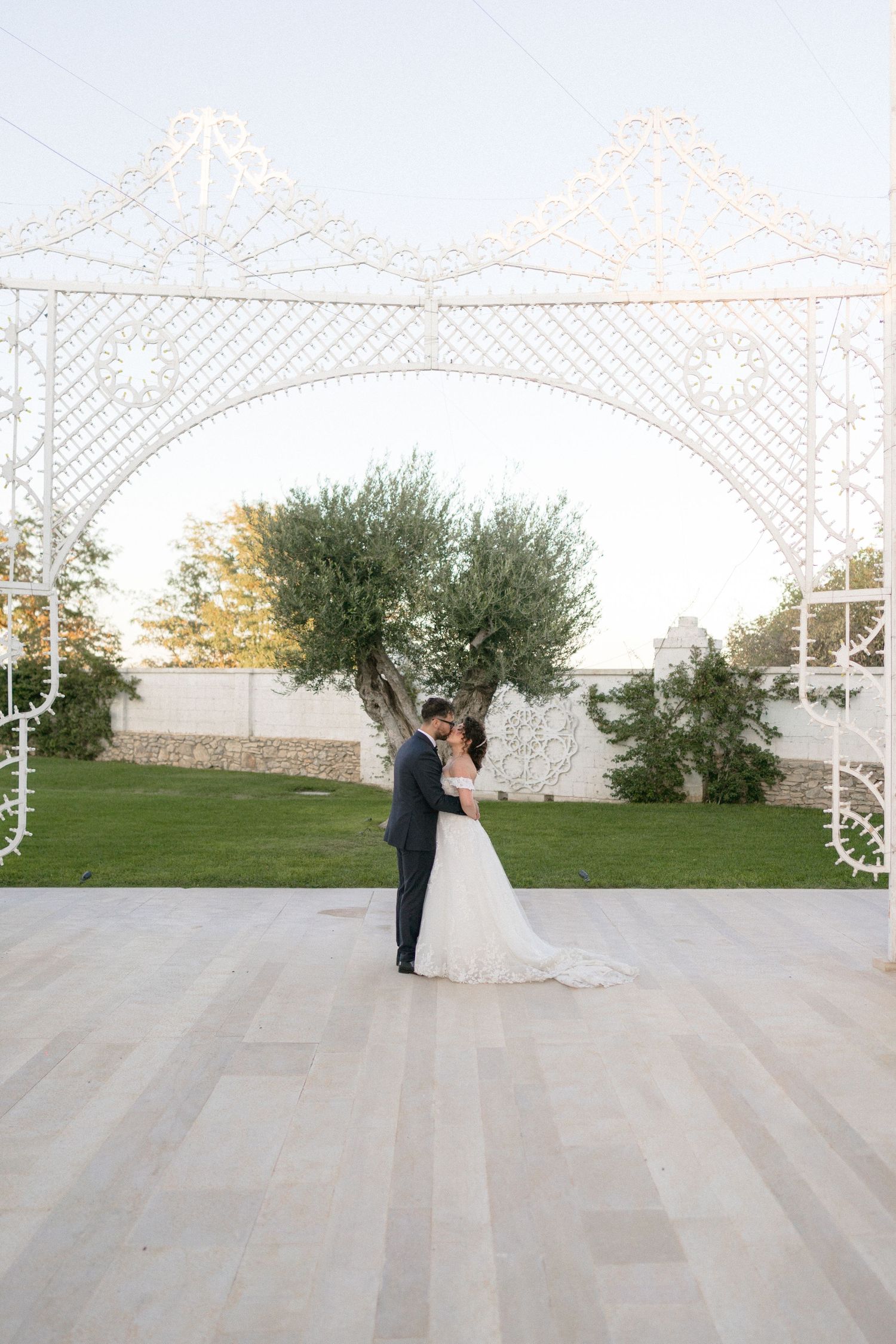 A bride and groom share a romantic kiss under a decorative white ornamental arch at golden hour outdoors.