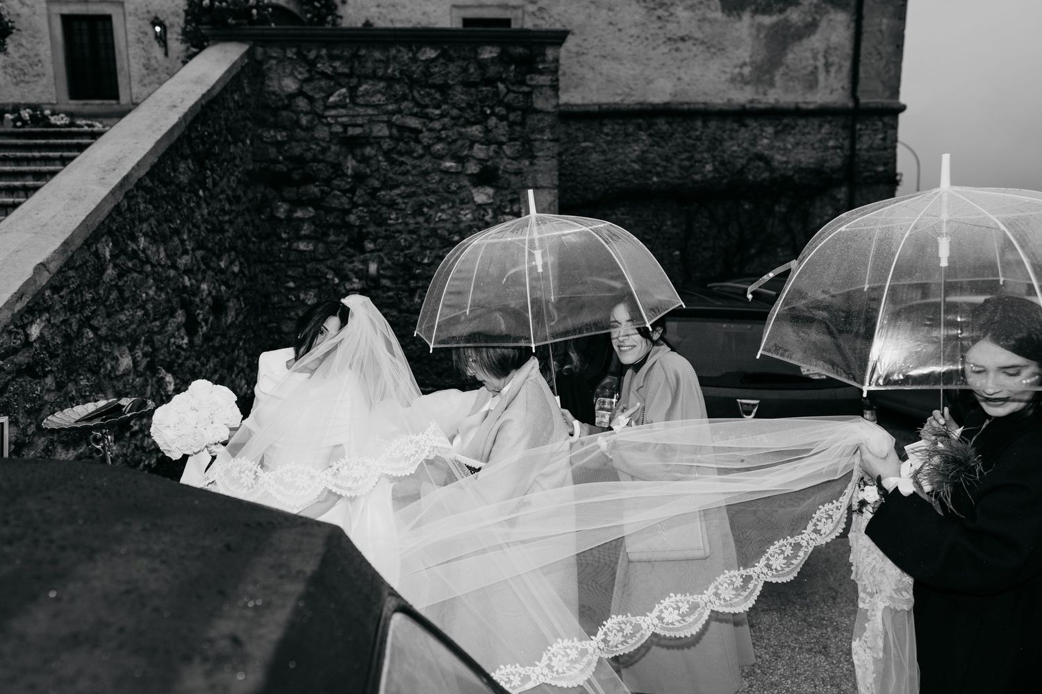 Black and white photo of bride and guests with umbrellas outside on a rainy wedding day.