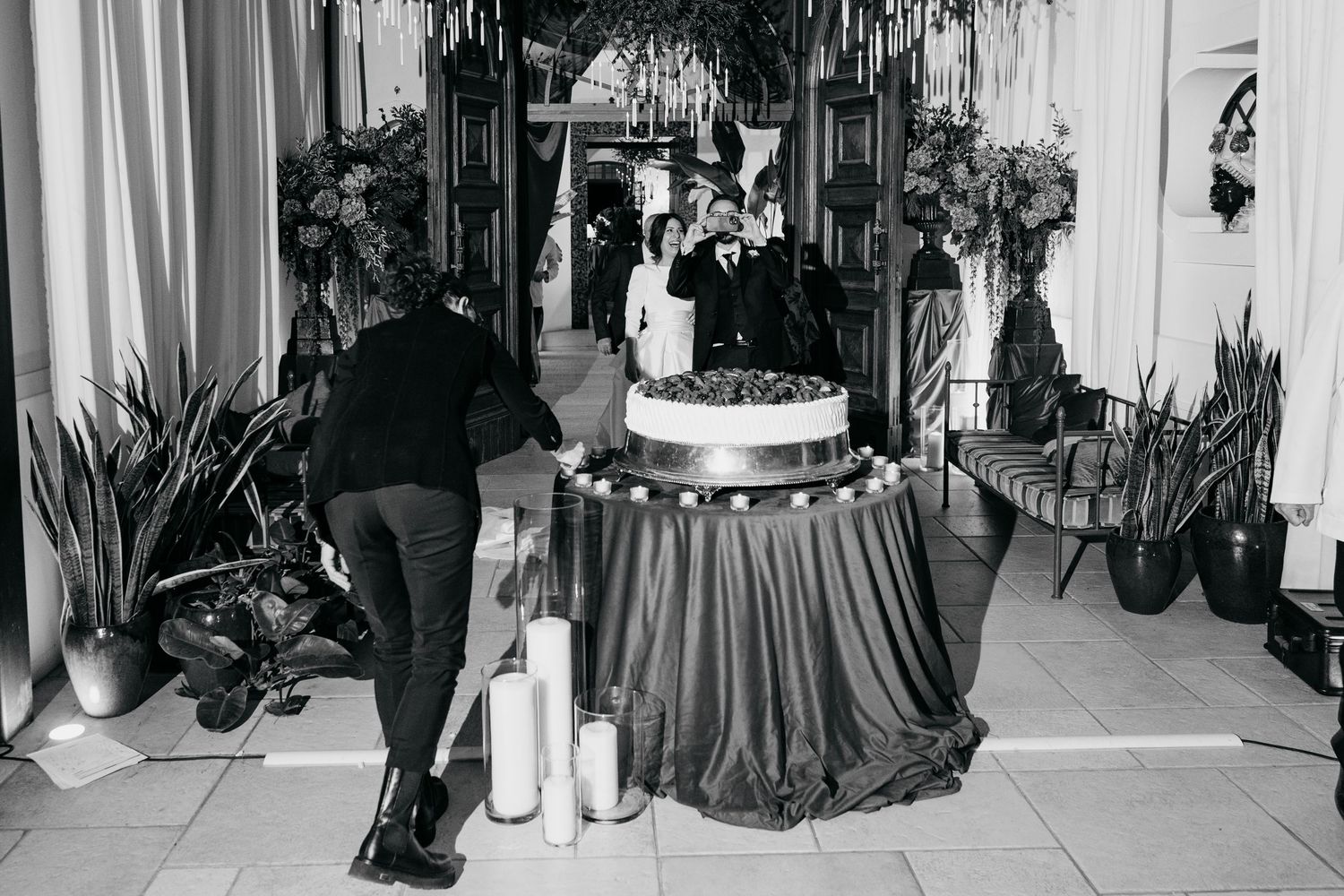 Black and white wedding reception scene with couple at tiered cake table surrounded by tall florals.
