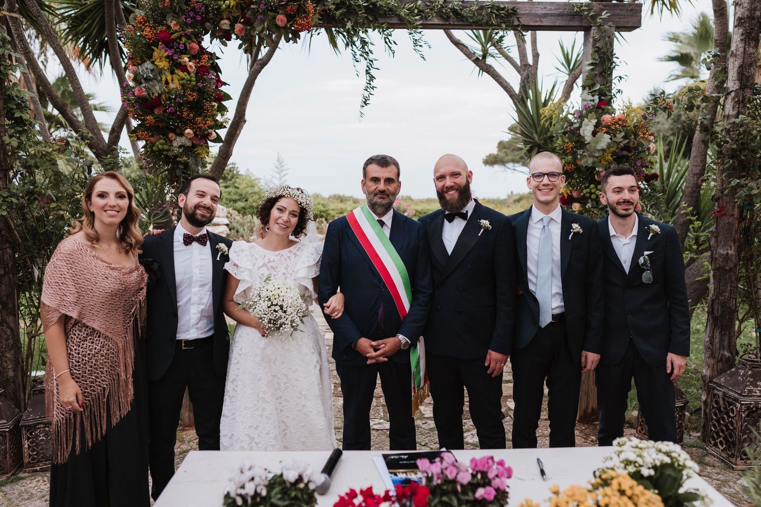 Wedding party poses together under floral arch at outdoor ceremony venue.