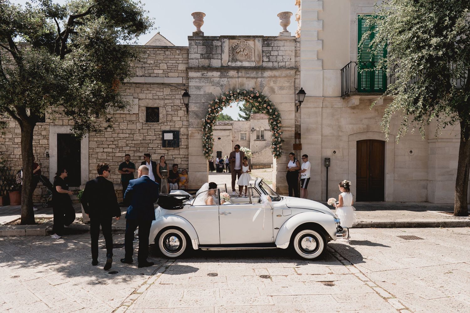 Classic white Volkswagen Beetle parked in front of a historic stone archway.