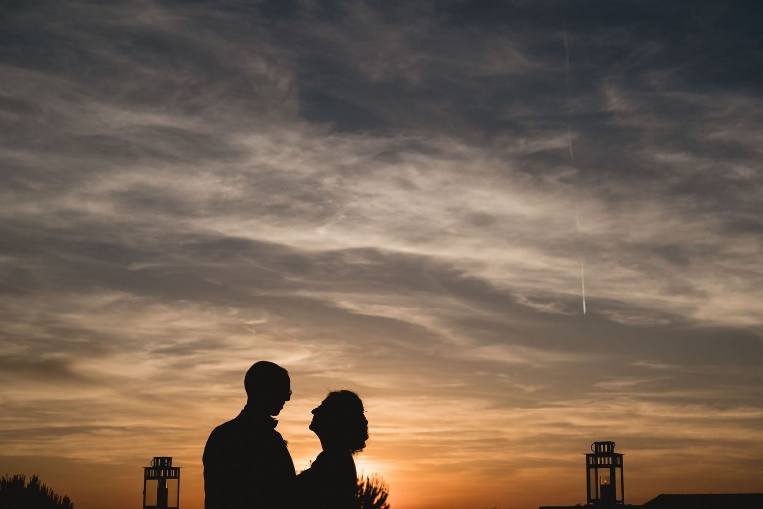 Silhouetted couple embracing against dramatic sunset sky with architectural elements in background.