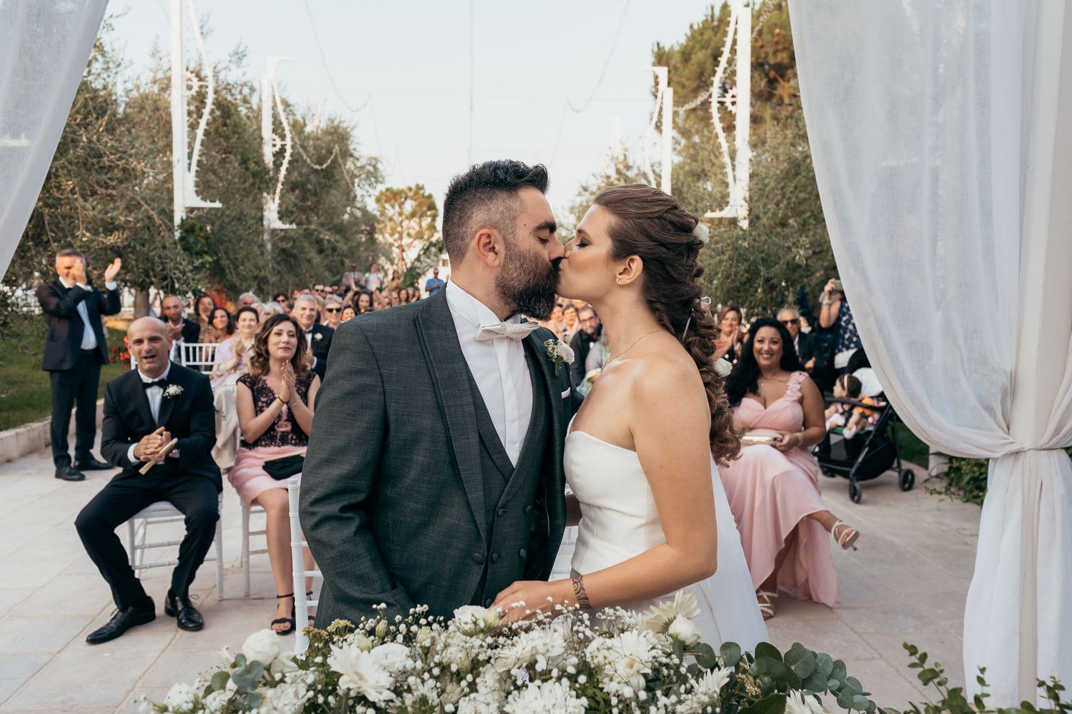 A tender moment shared between newlyweds under white drapes with wedding guests in background.