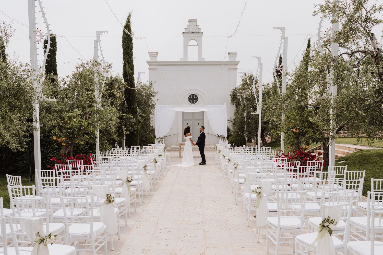 A white chapel at the end of an aisle lined with chairs and cypress trees for an outdoor wedding.