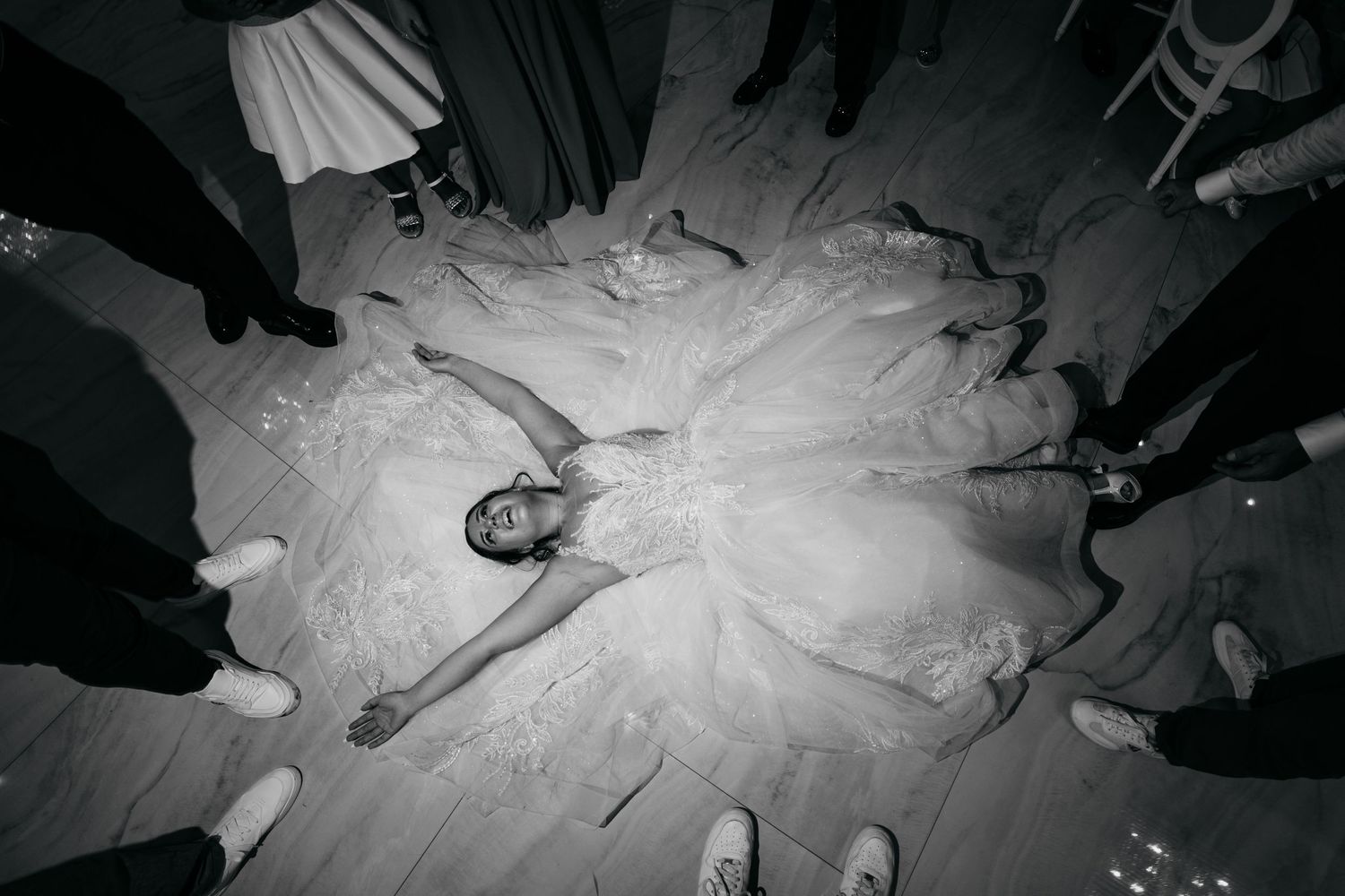Overhead view of a bride in white dress laying on floor surrounded by standing guests at wedding reception.