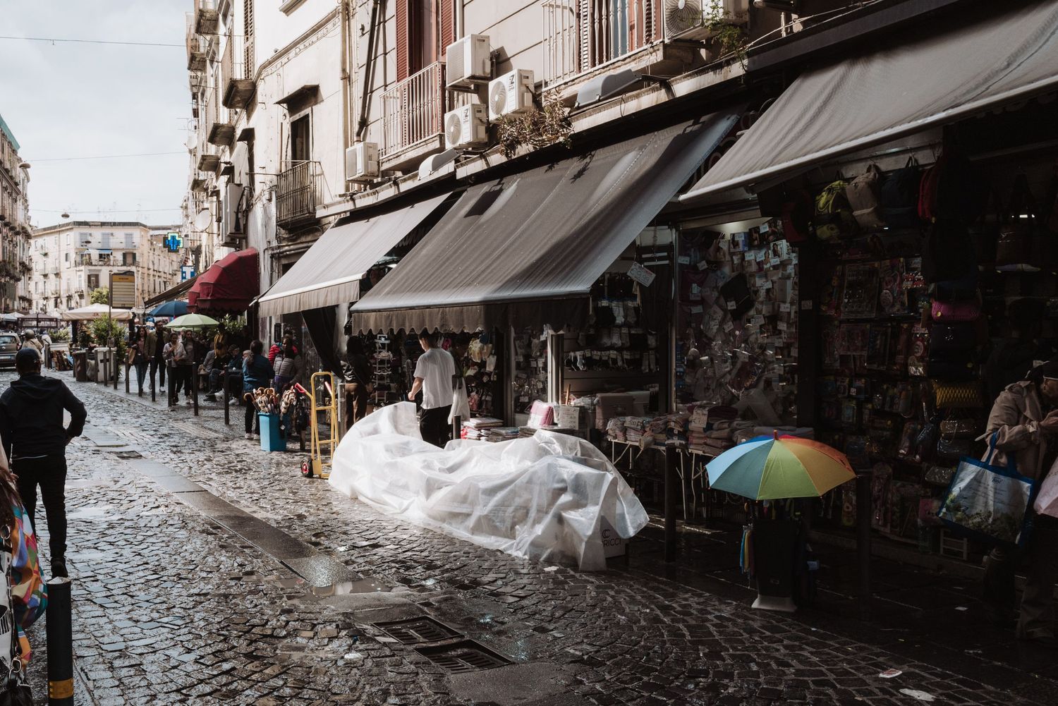 People walking along a rainy street with shops and awnings in Naples, Italy on a wet cobblestone road.