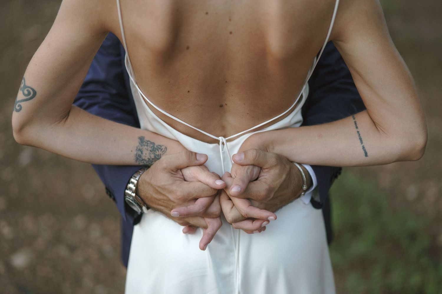 A person in a white dress holds their hands behind their back, showing visible tattoos on their arms.