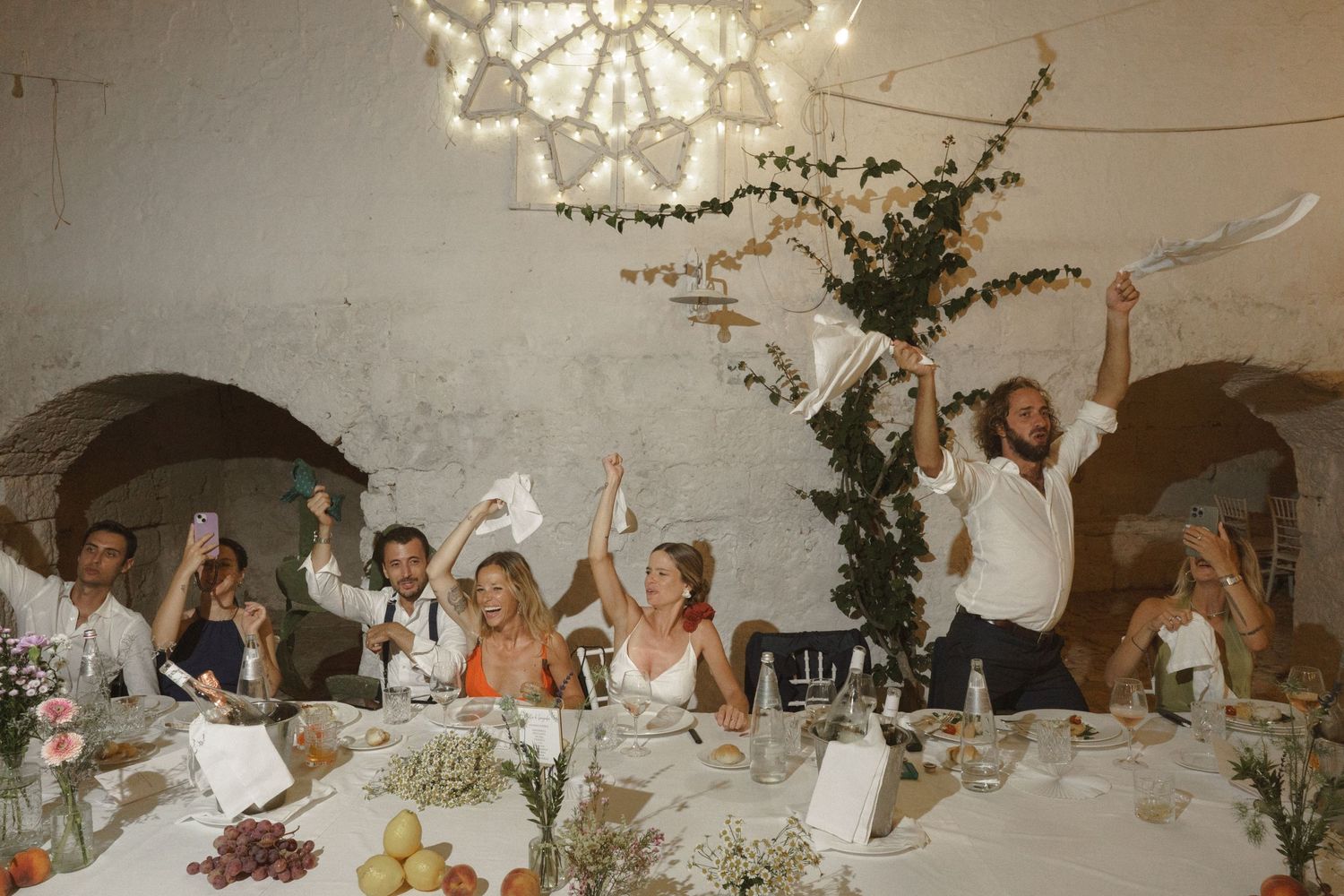 Wedding guests celebrate with raised arms at a candlelit reception table under an ornate chandelier and stone arches.