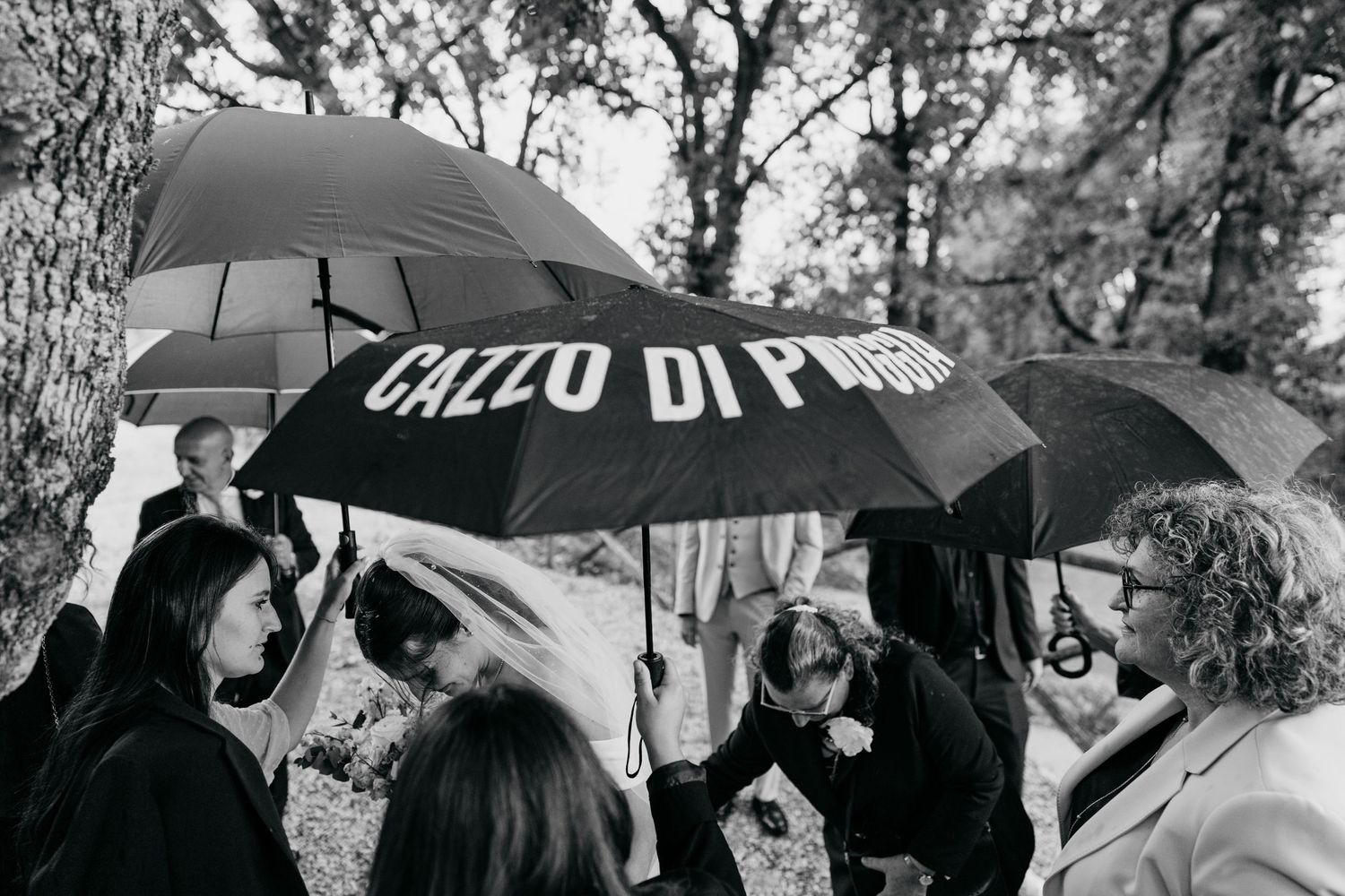 A group gathers under a black umbrella with text during an outdoor celebration.