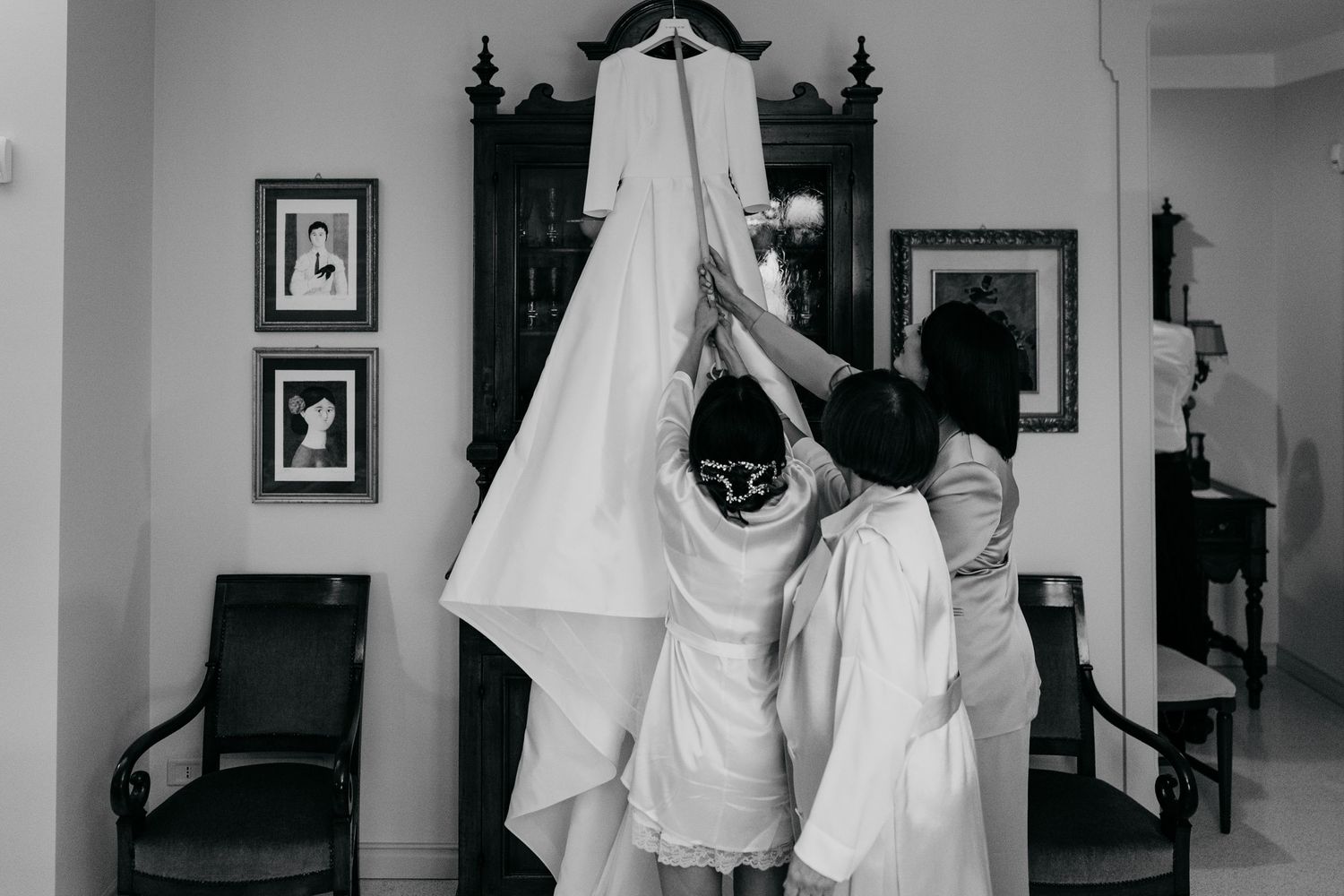 Two women in robes reach up to touch a hanging white wedding dress in a classic room with framed photos.