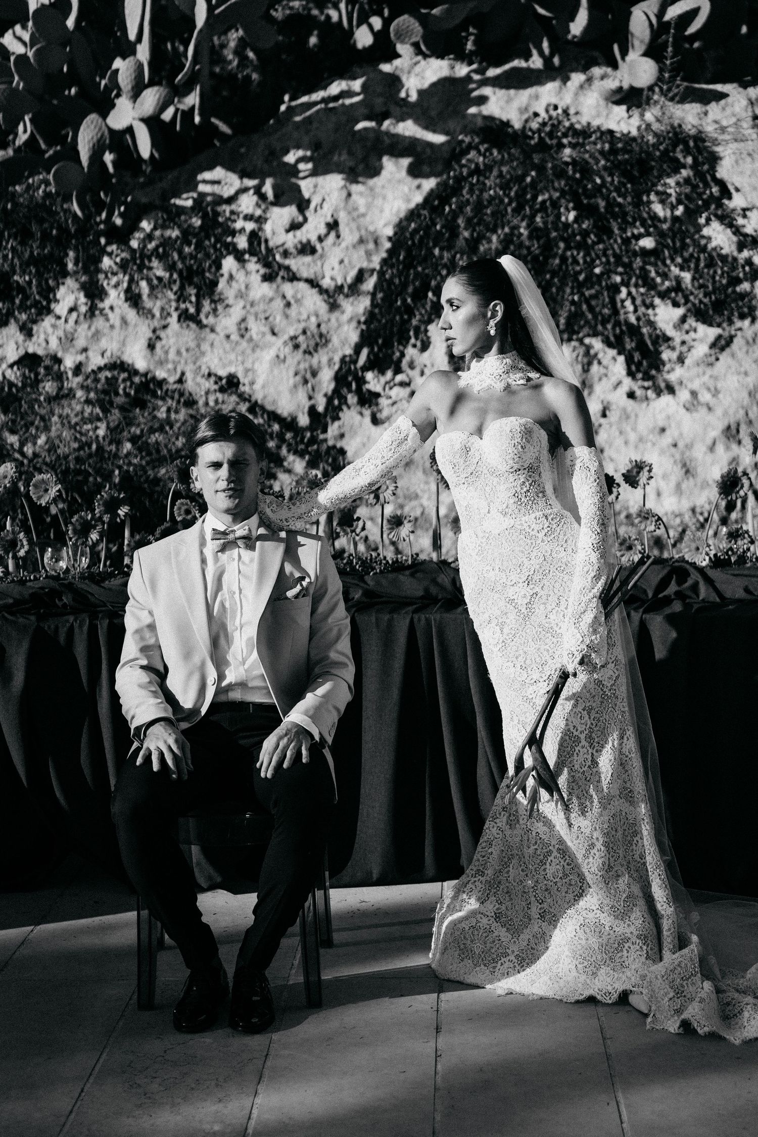 Black and white wedding portrait of a seated groom in a white dinner jacket with the bride in a lace gown standing beside him.