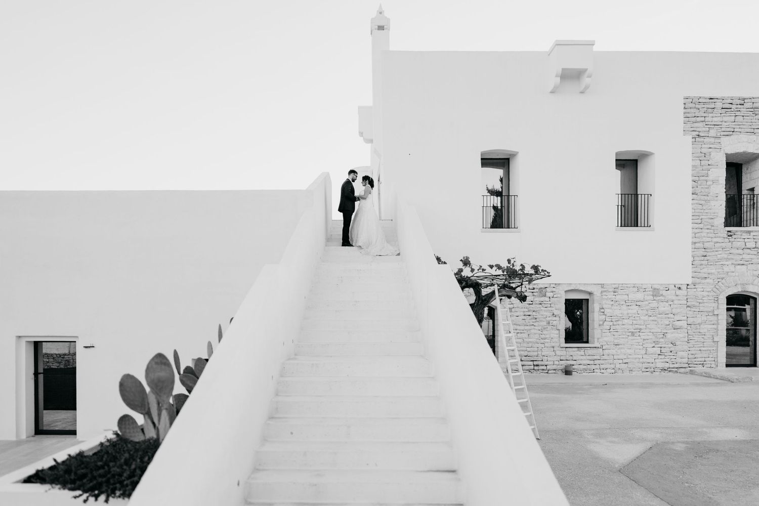 A bride and groom stand atop white stone stairs at a minimalist whitewashed Mediterranean venue in black and white.