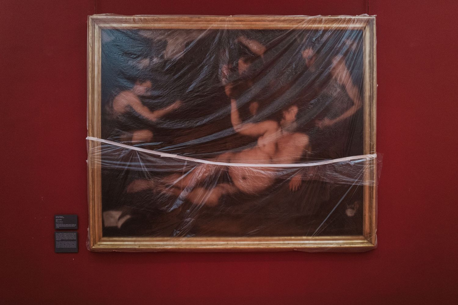 A framed artwork covered in protective plastic wrap hangs on a dark red wall next to a small black box.