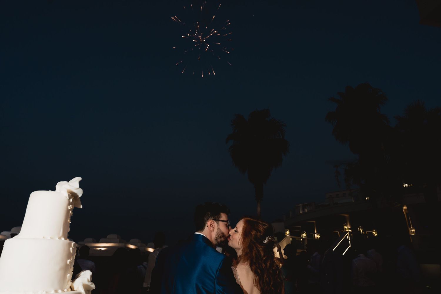 Couple kissing under fireworks during their nighttime wedding reception.