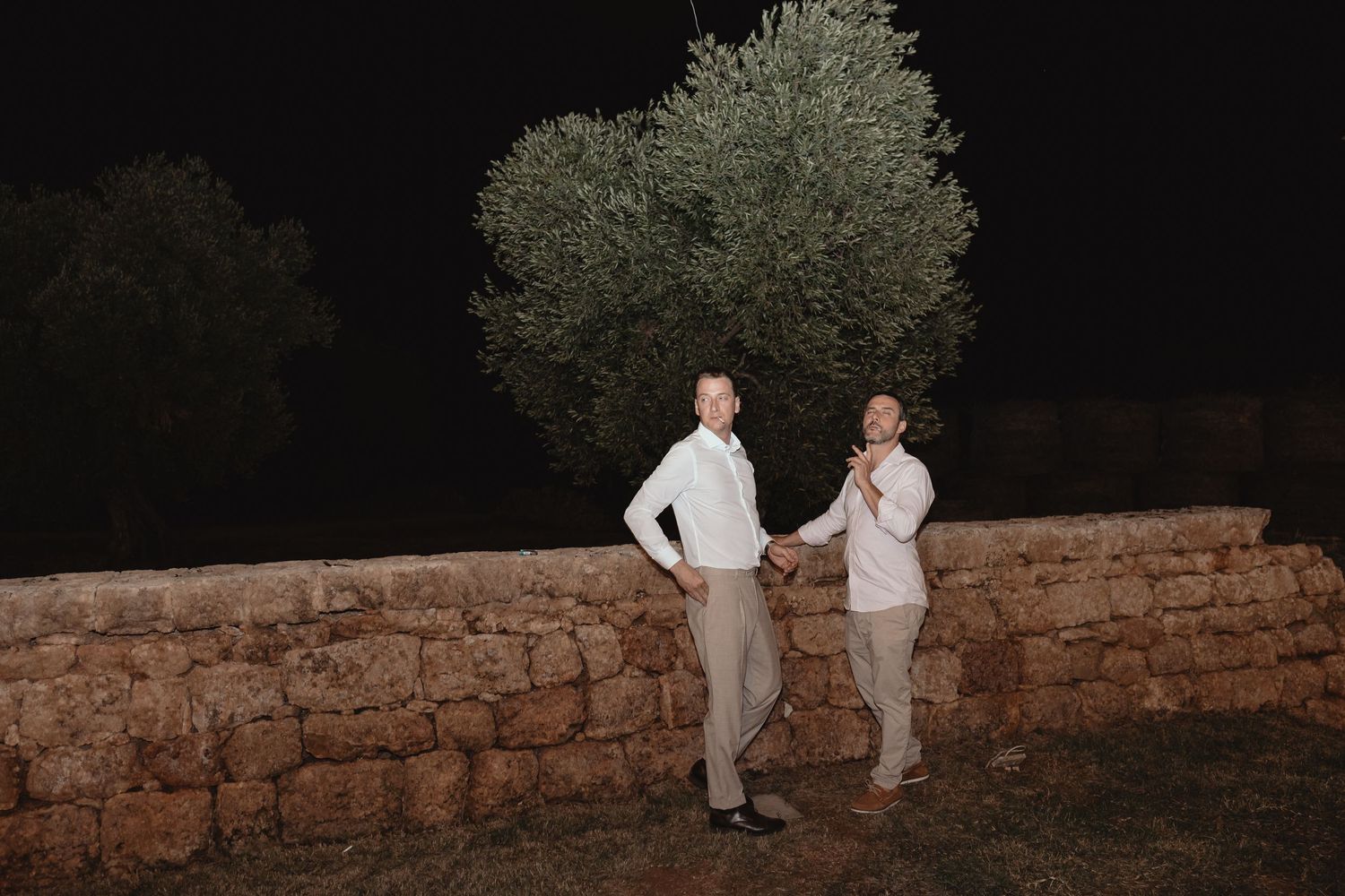 Two people in white outfits stand on a rustic stone wall in a garden setting at dusk.