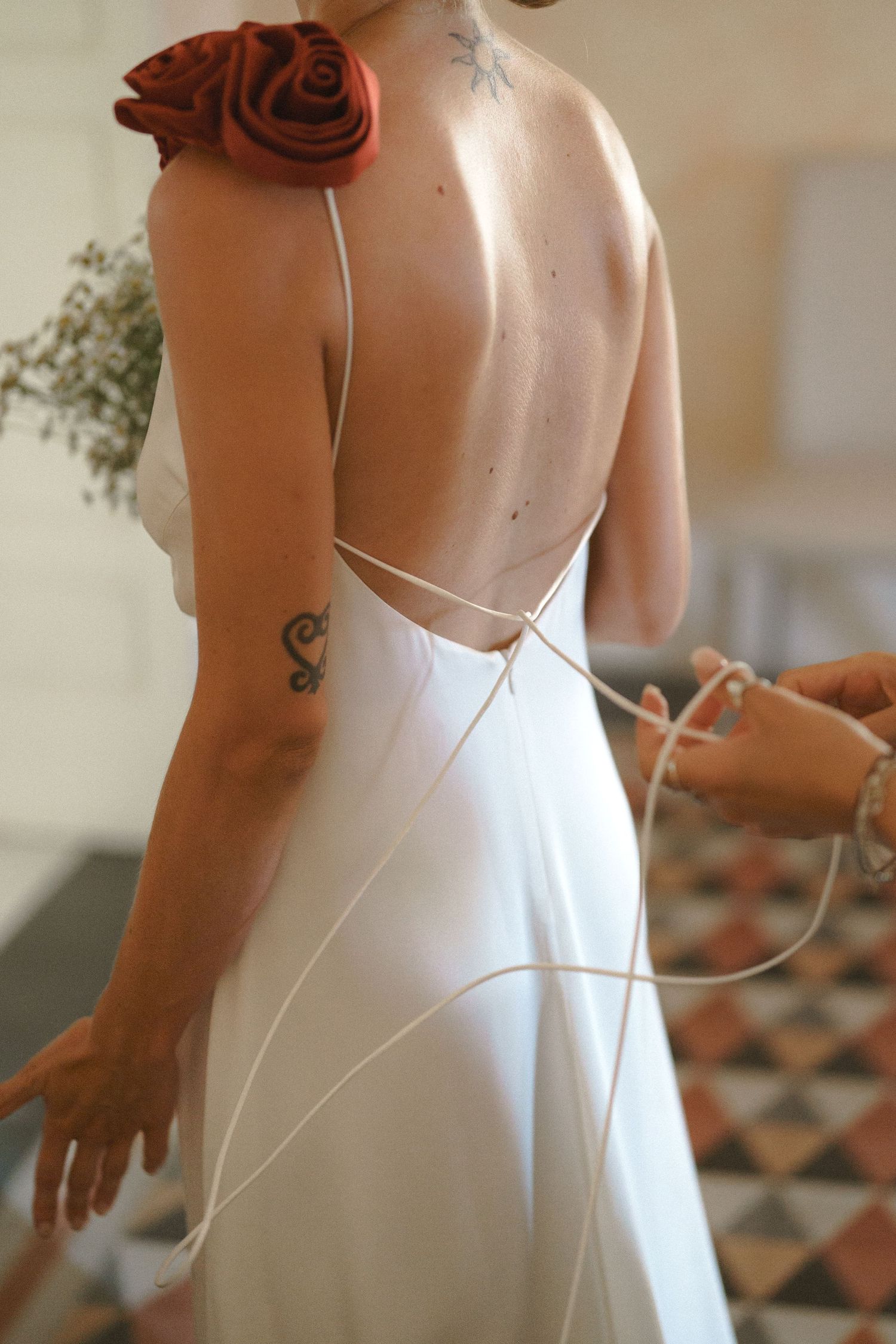 Elegant backless white wedding dress with spaghetti straps and a red rose accent shows a small arm tattoo.