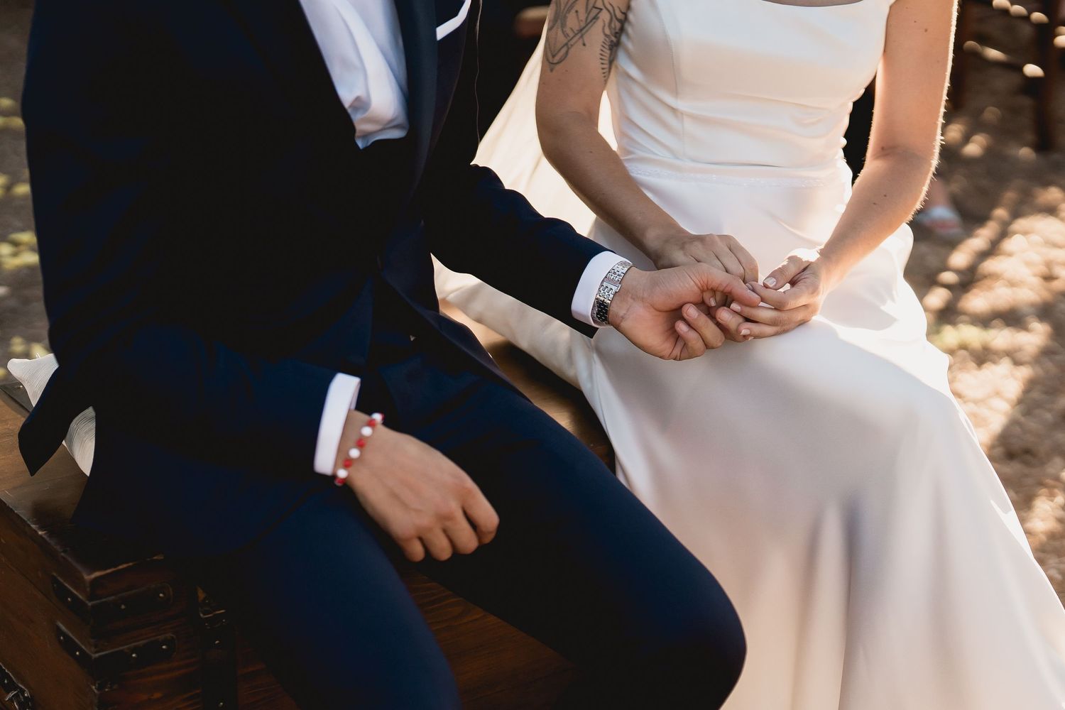 Cropped wedding photo showing seated couple in traditional navy suit and white gown holding hands in dappled sunlight.