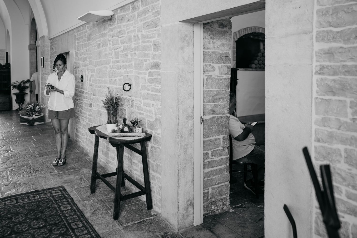 A black and white photograph showing a casual indoor scene with stone walls, a small table, and two people in separate spaces.