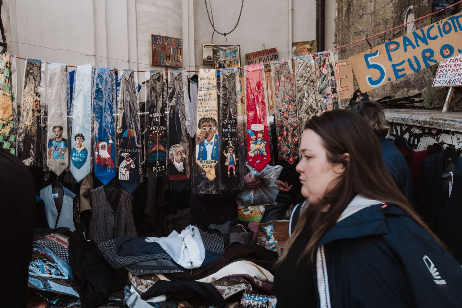 Person shopping at an outdoor market stall with various items displayed on a wall behind them.