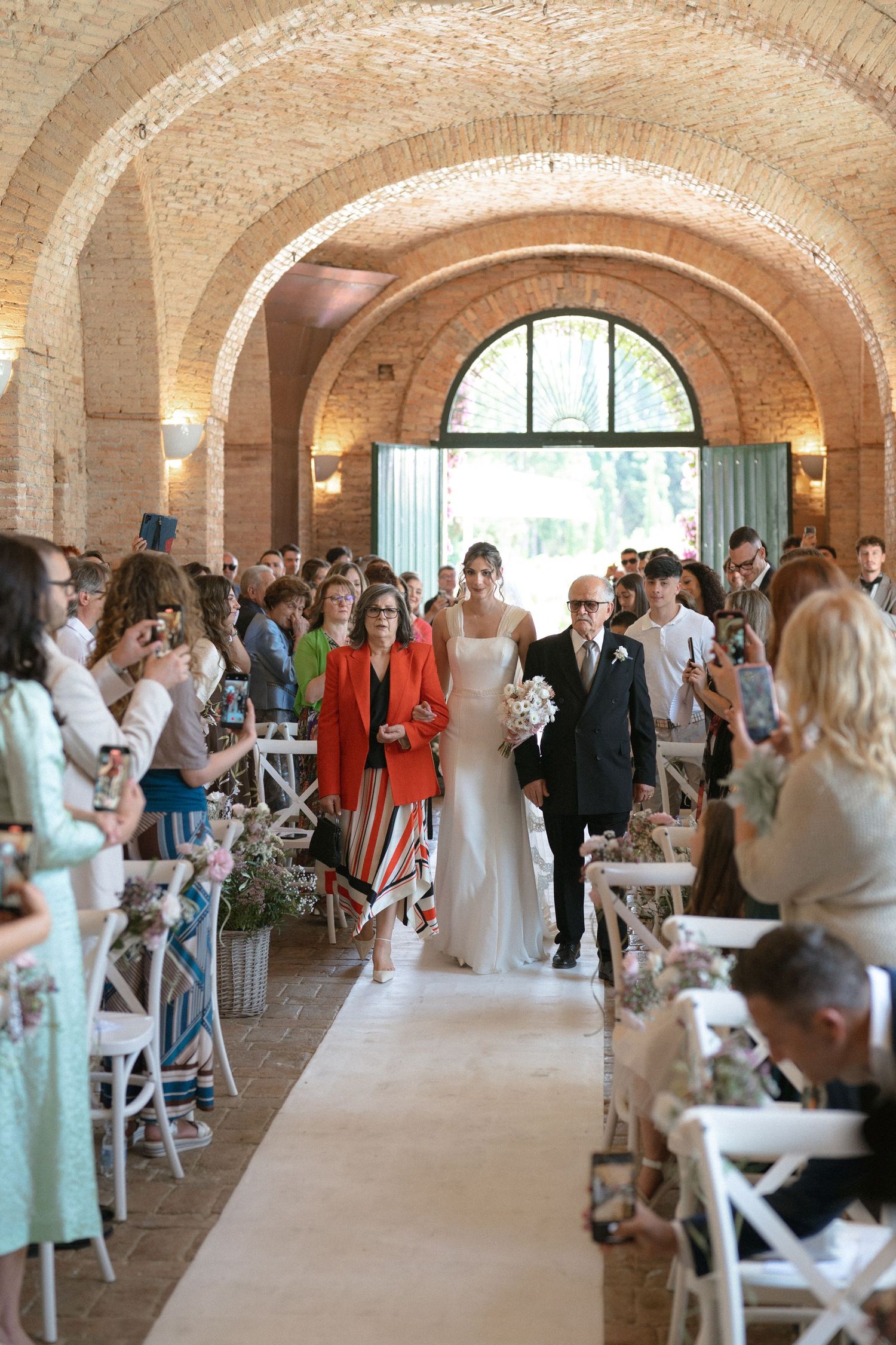 A wedding ceremony takes place in a beautiful brick-vaulted room with arched ceilings as guests watch from white chairs.