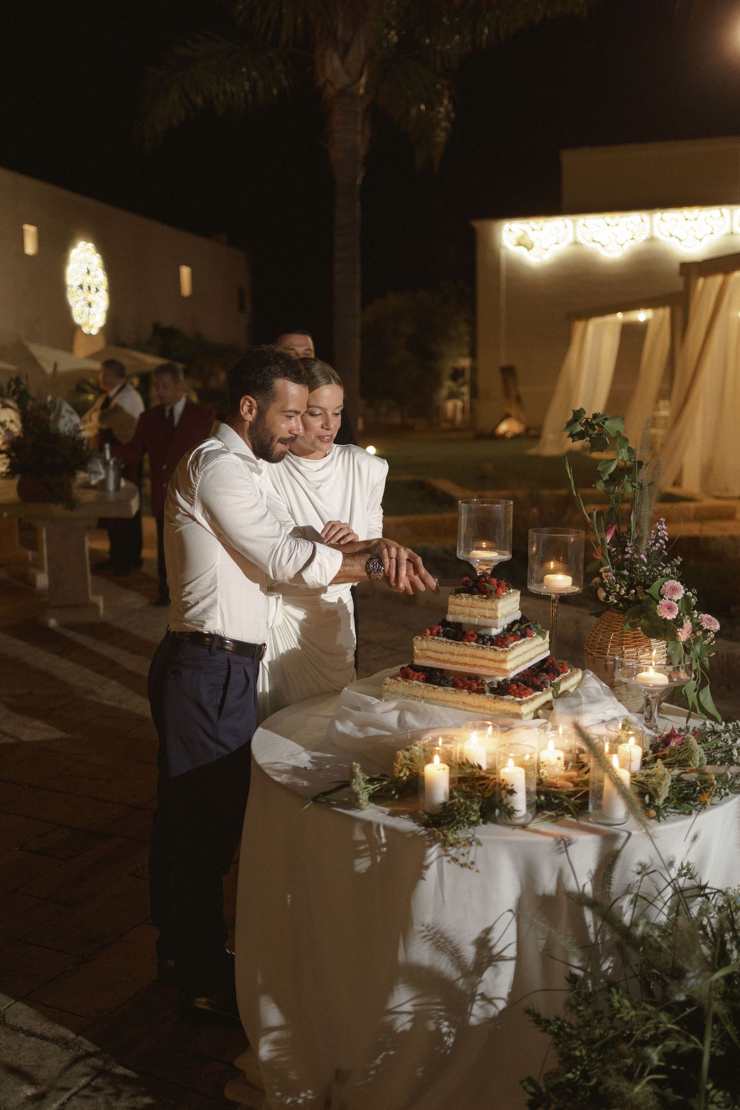 A couple cutting their wedding cake together at an elegantly decorated reception table with candlelight and greenery.