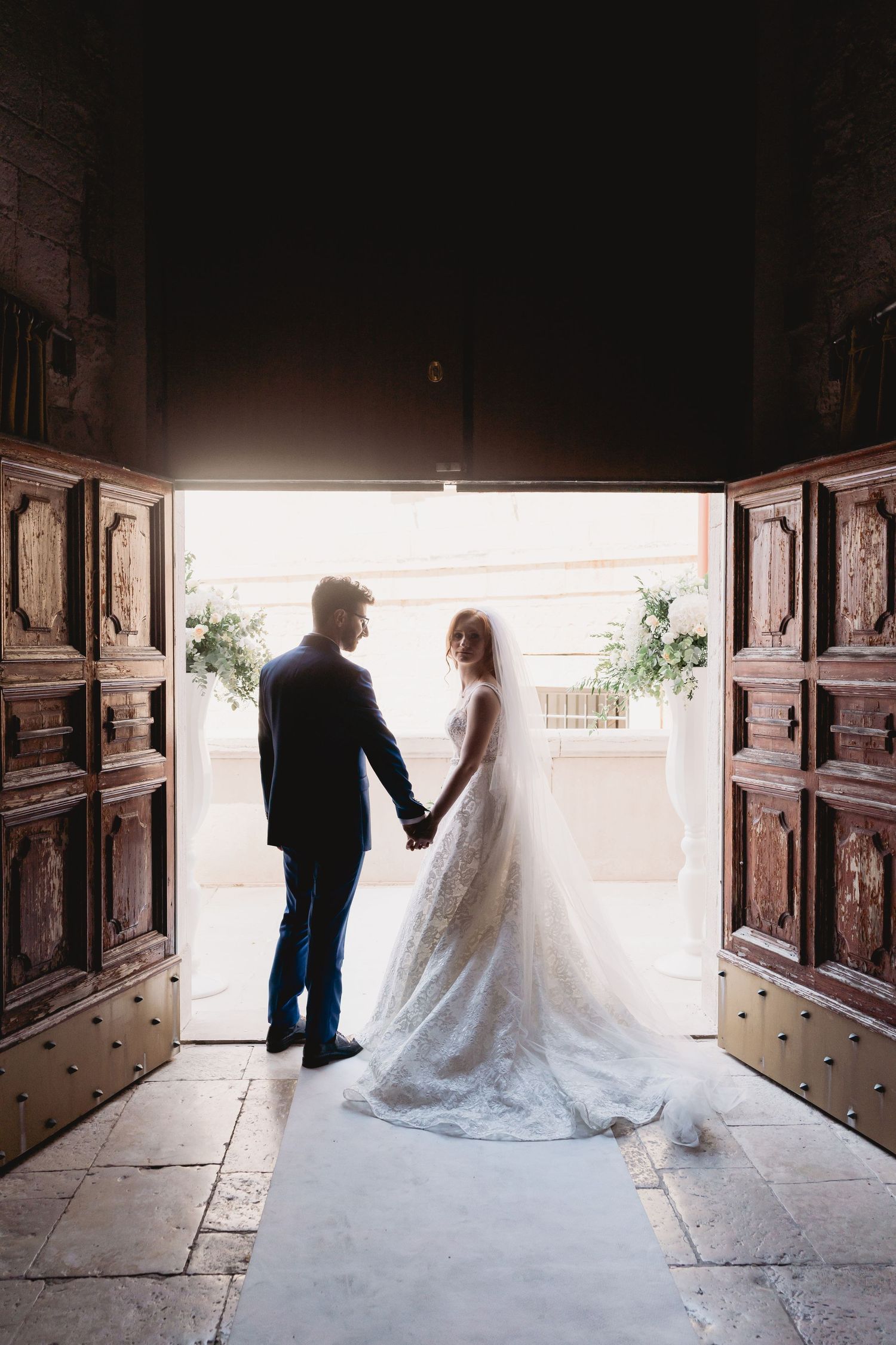Silhouetted wedding couple holds hands while standing in a grand doorway with ornate wooden doors.
