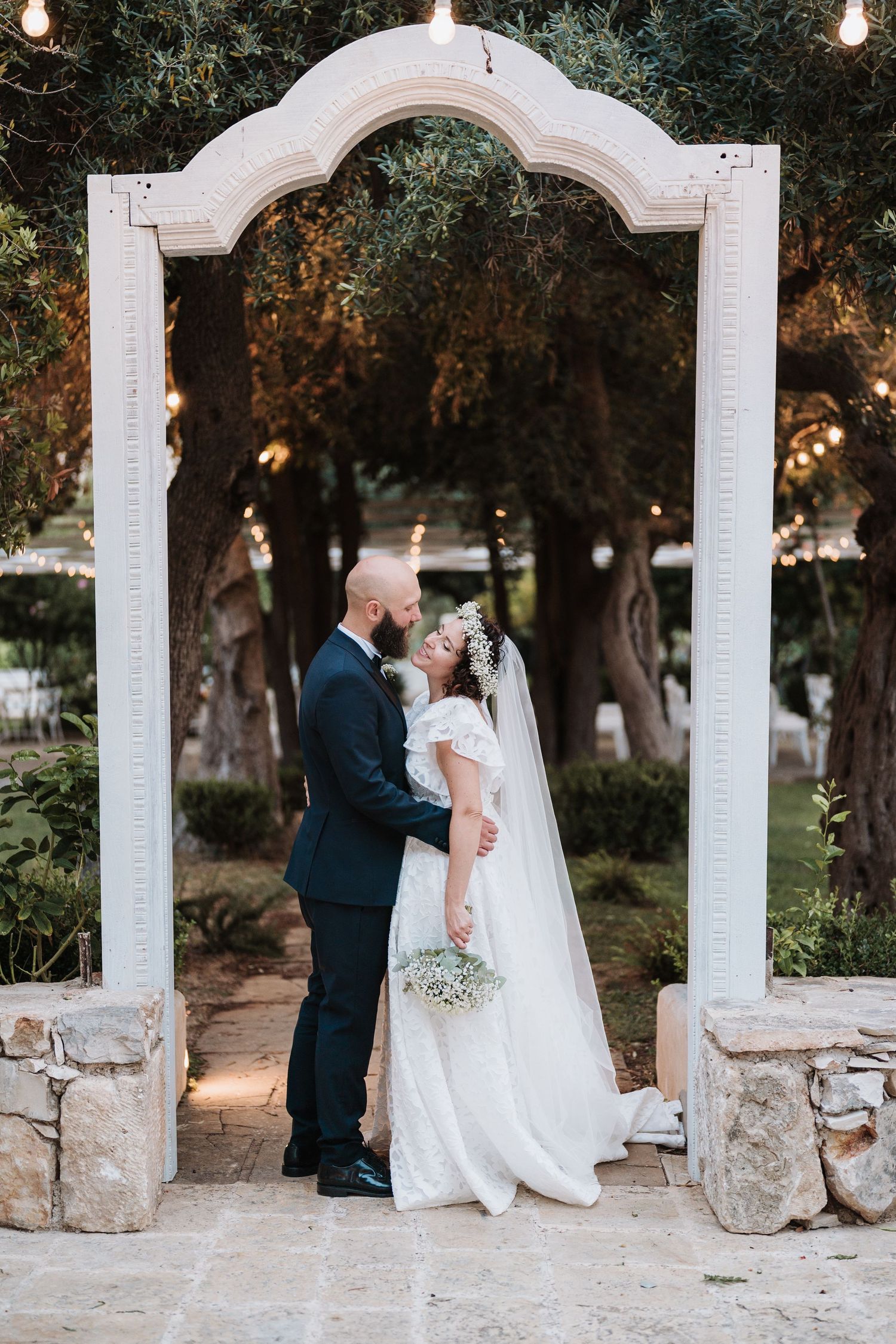 A romantic wedding portrait under a white decorative arch with garden lights in the background.