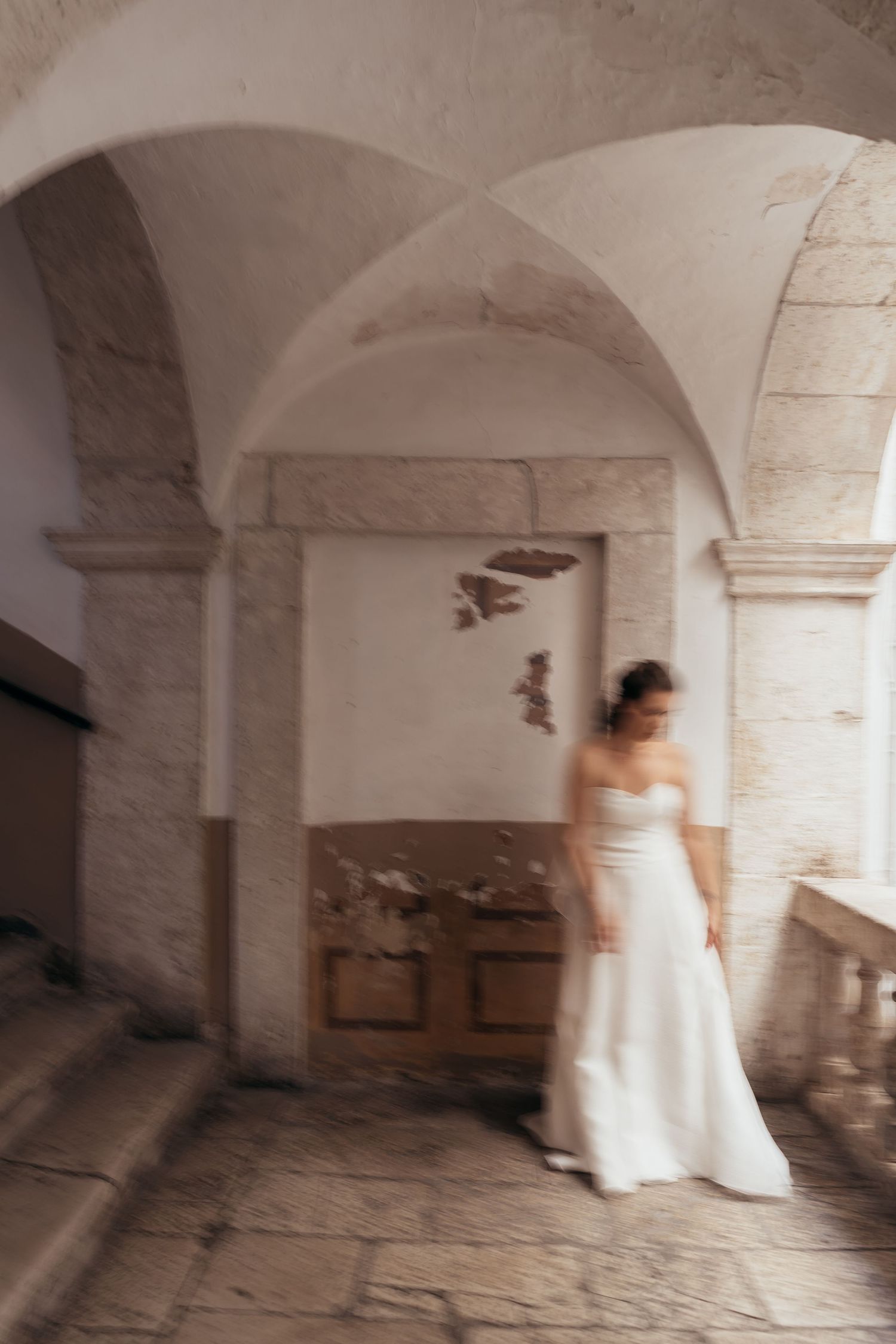 Artistic motion blur photograph of a bride in white dress standing under stone archways.