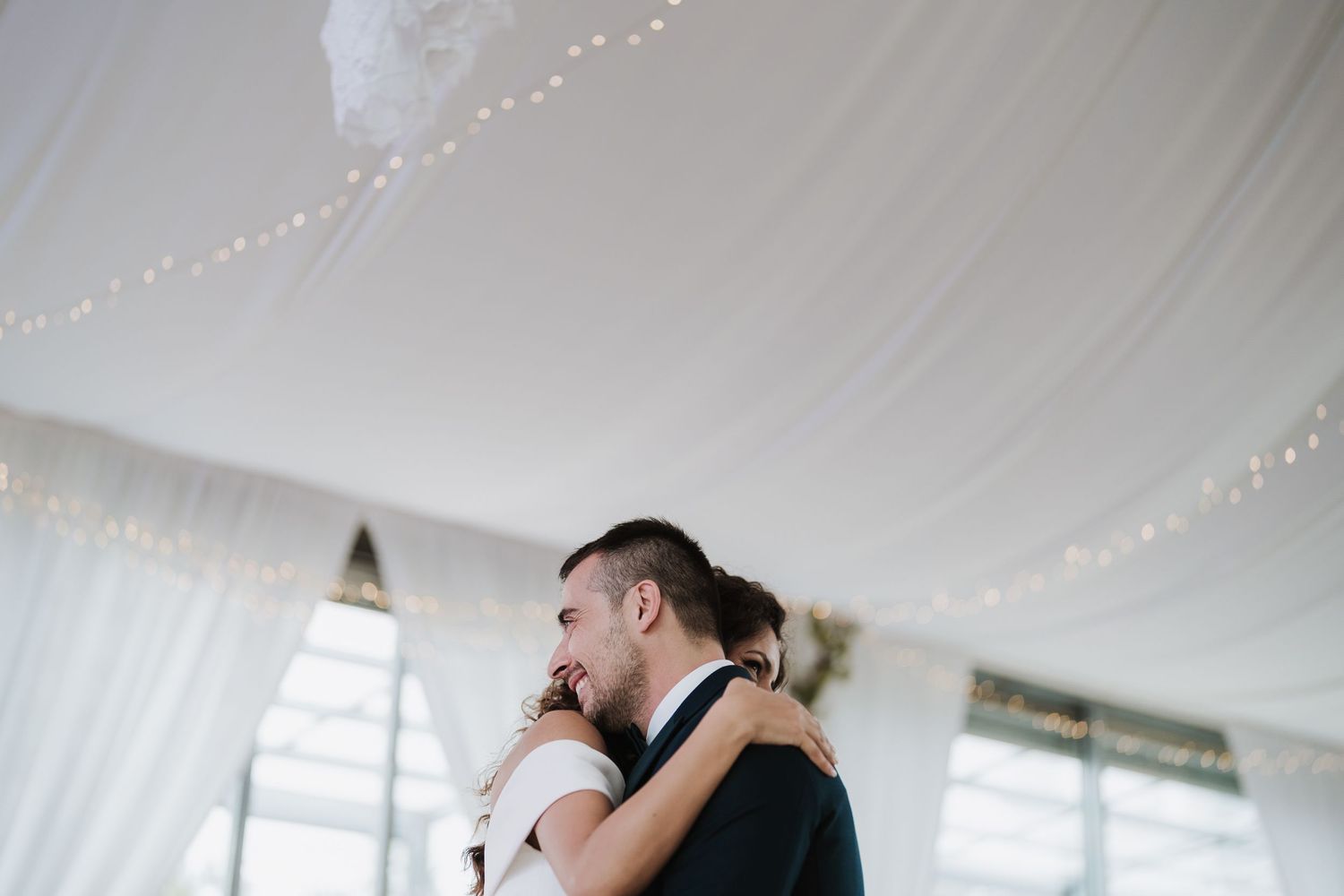 Couple embracing under white tent with string lights at wedding reception.