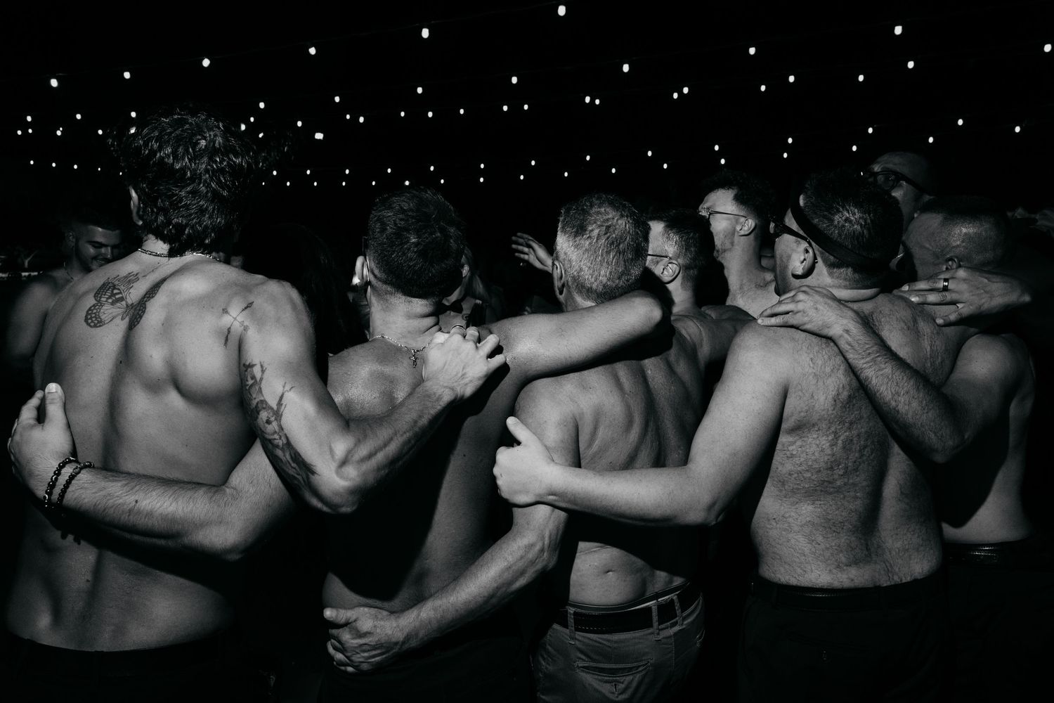 Shirtless wedding guests dancing close together under string lights at night.