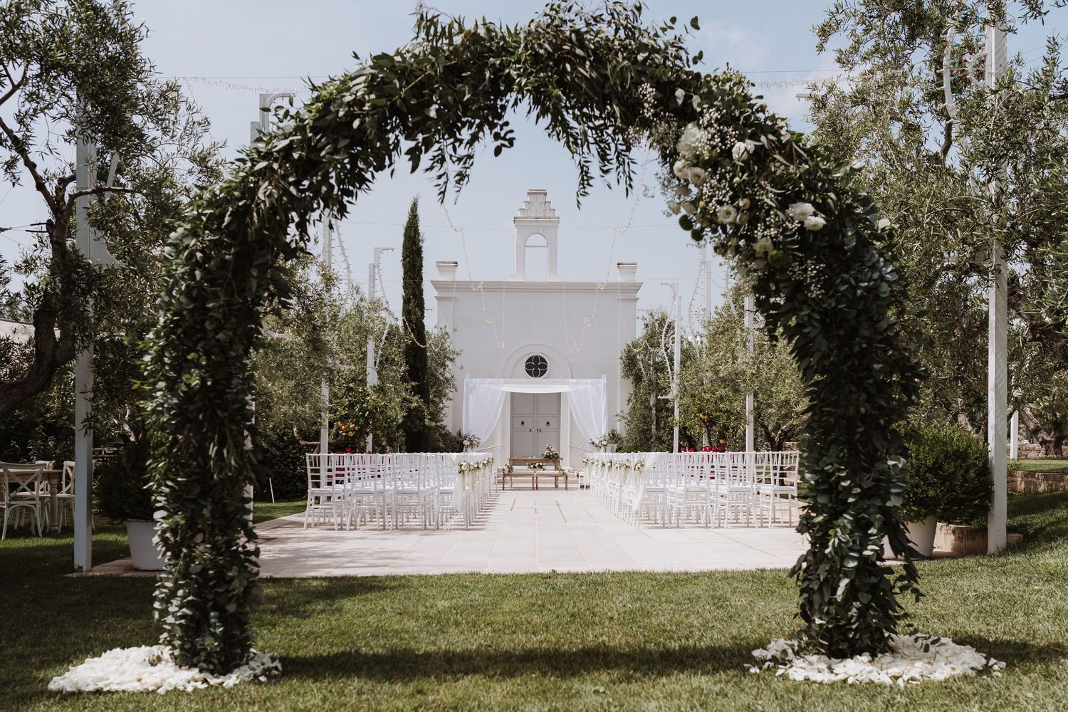 Wedding ceremony setup with floral arch and white chairs facing a white chapel.