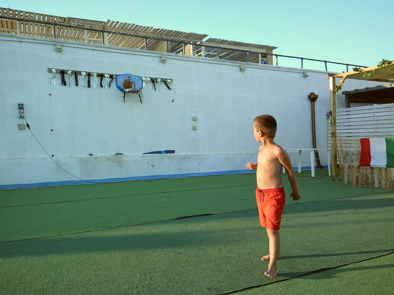 Child in red shorts walking on green artificial turf near a white wall.