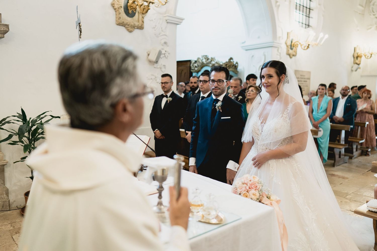 A traditional Catholic wedding ceremony takes place in an ornate white church with arched ceilings and chandeliers.