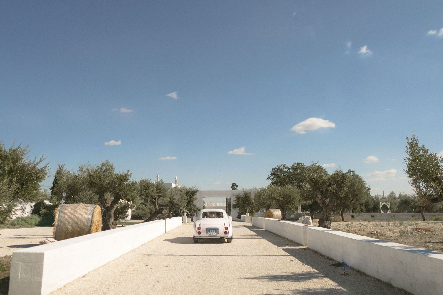 Vintage white Fiat 500 driving down a tree-lined gravel path at a rustic wedding venue.