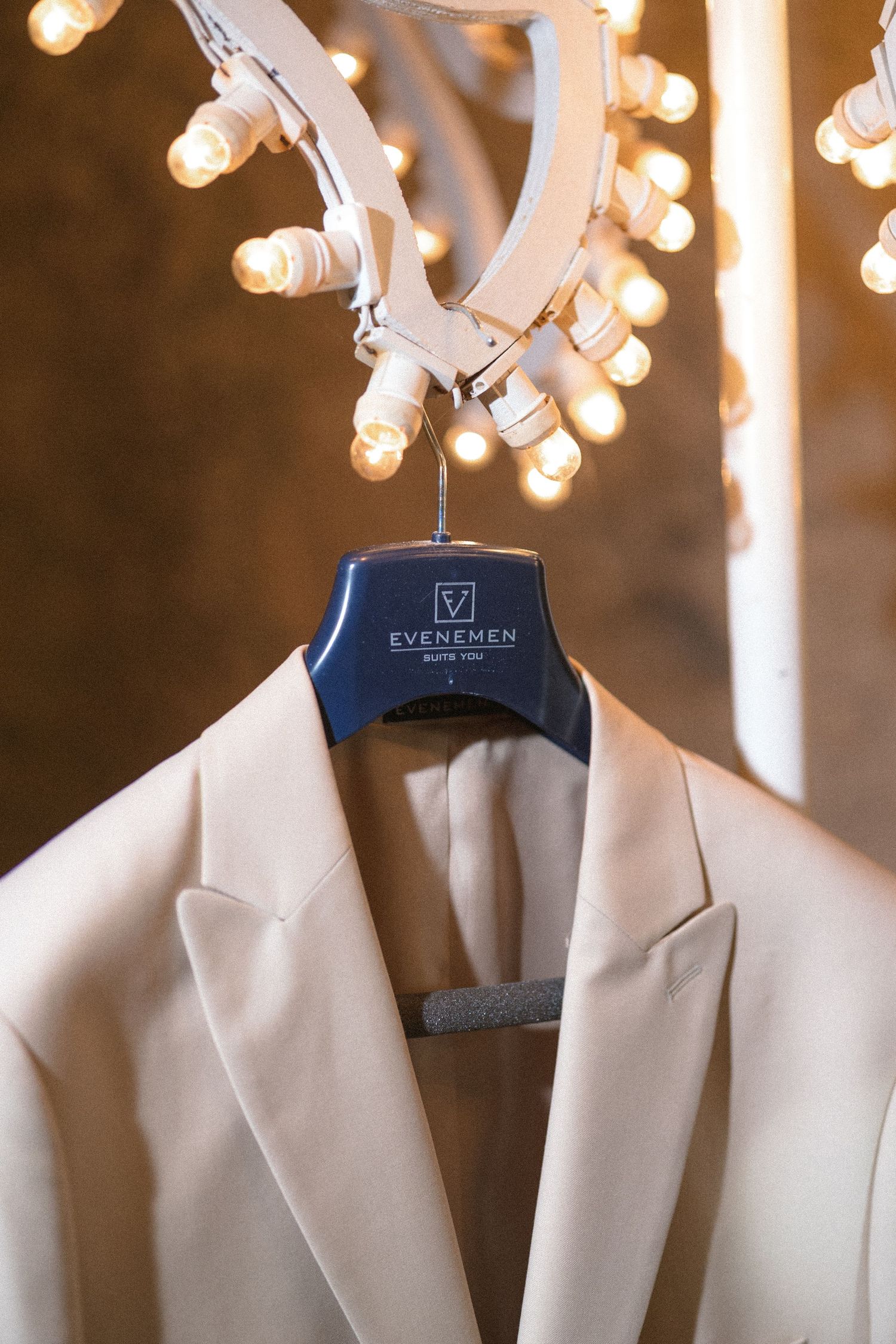A cream double-breasted blazer hangs on a navy Evenemen hanger beneath a chandelier of glowing bare bulbs.