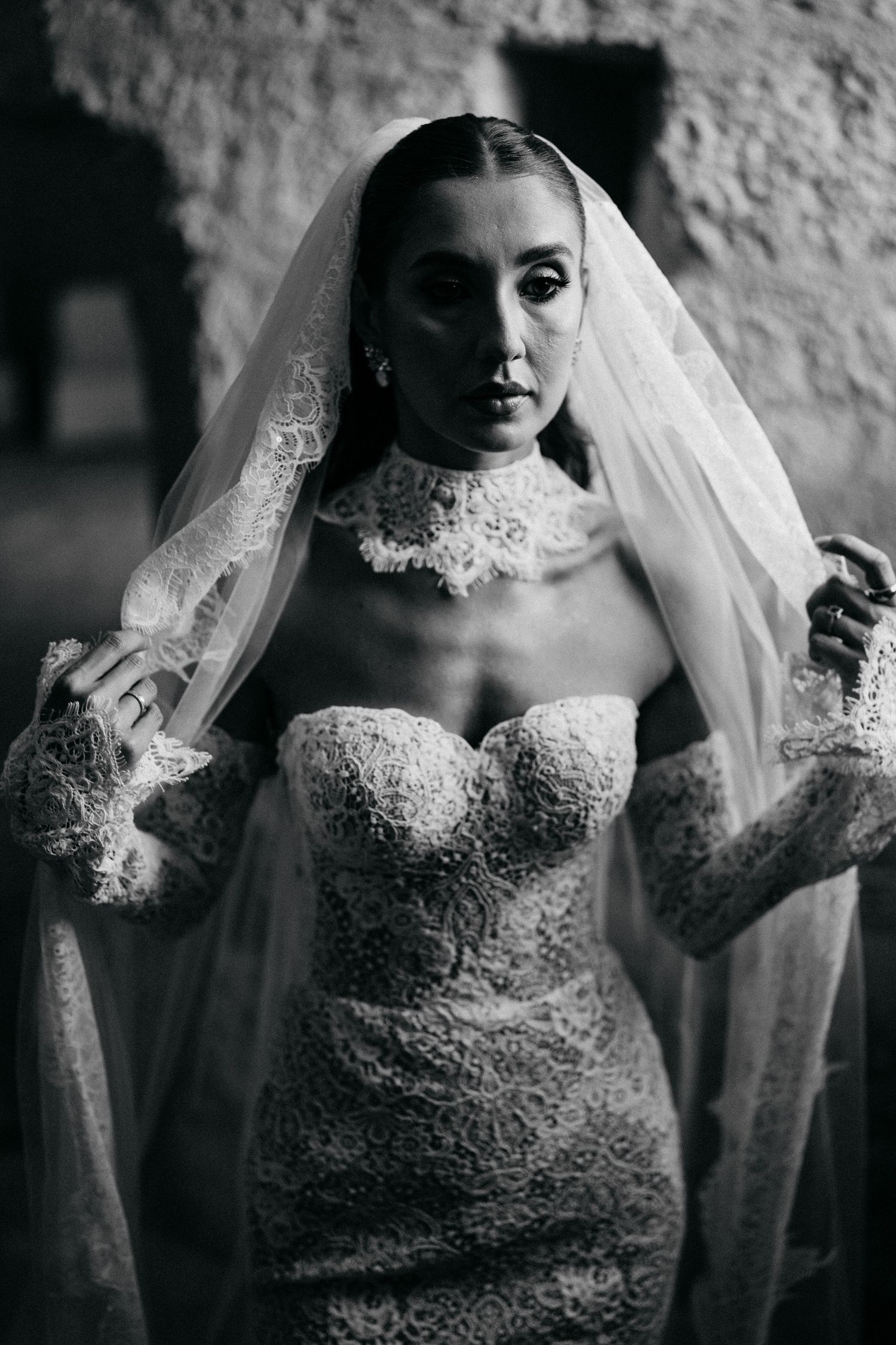 Black and white portrait of a bride in a strapless lace gown with high collar and lace veil against a rustic stone wall.