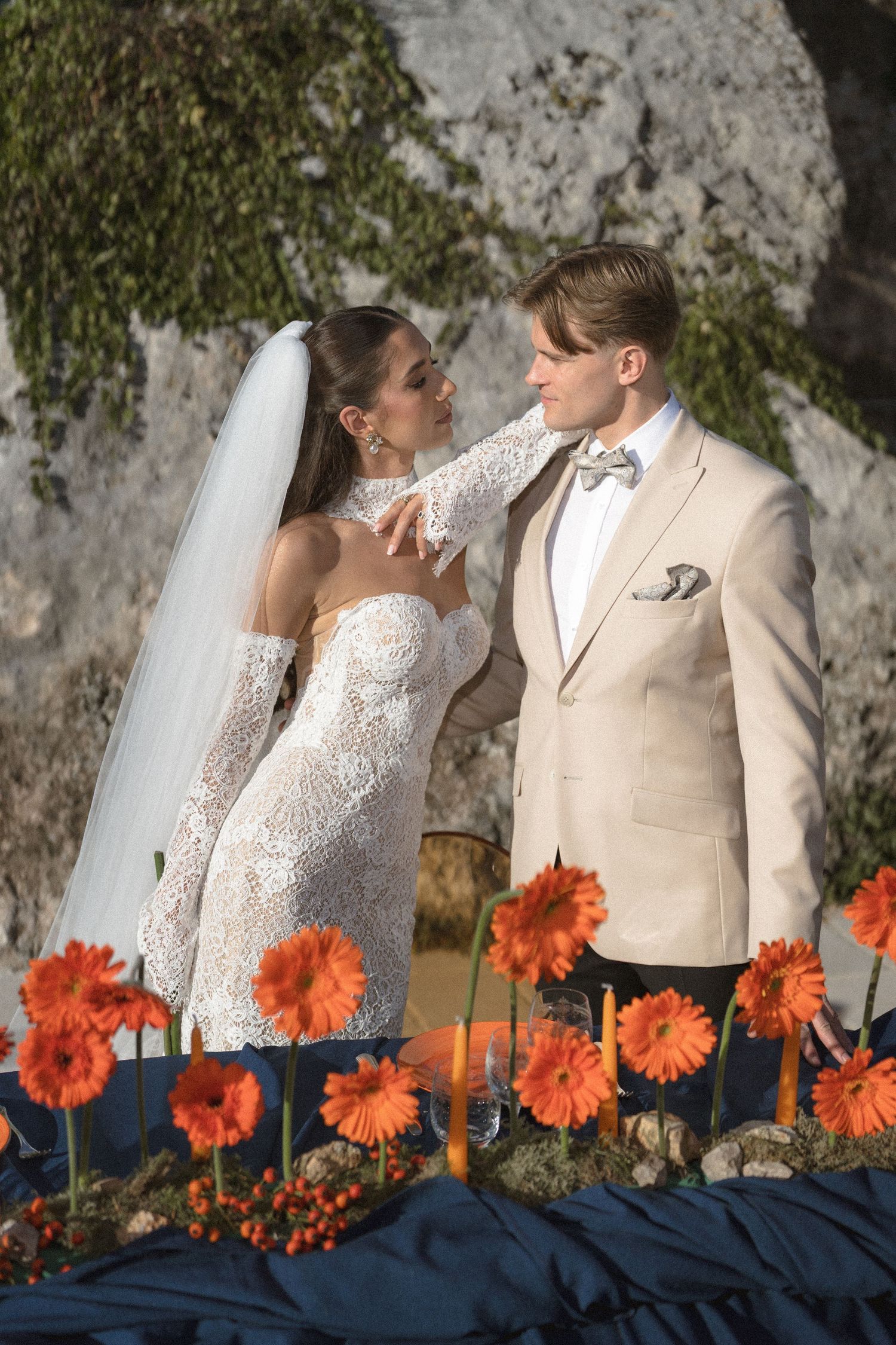 Bride in a lace wedding gown and groom in a beige suit with bow tie embrace beside orange gerbera daisies at an outdoor ceremony.