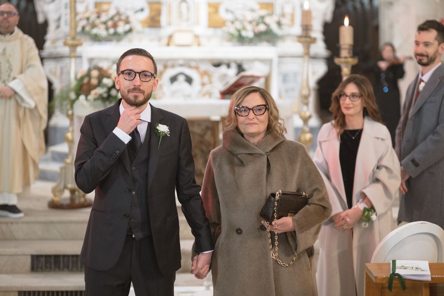 Groom and older woman walk down church aisle during wedding ceremony, altar visible in background.