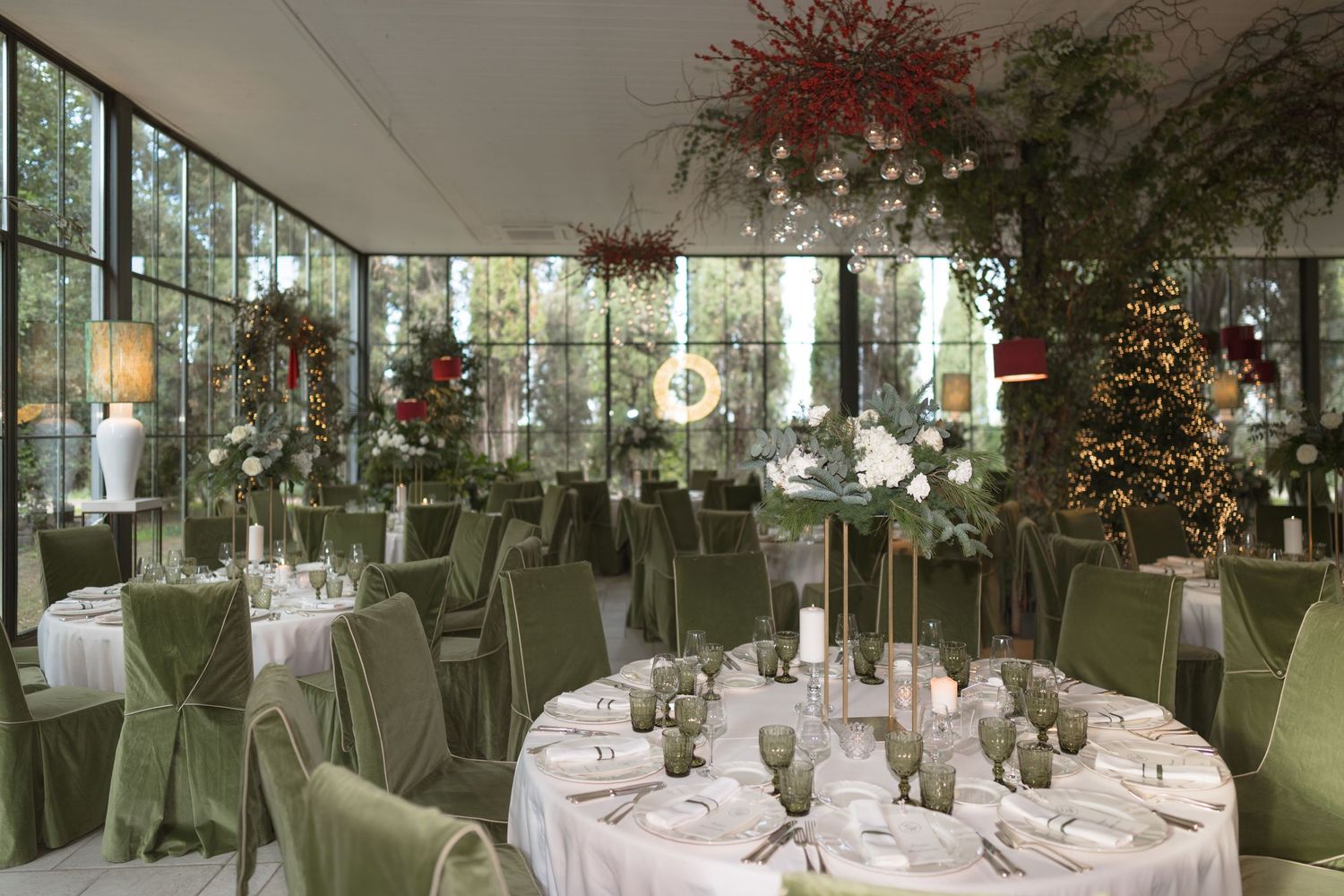 Elegant wedding reception venue with green velvet chairs, round tables, lush floral arrangements and large windows.