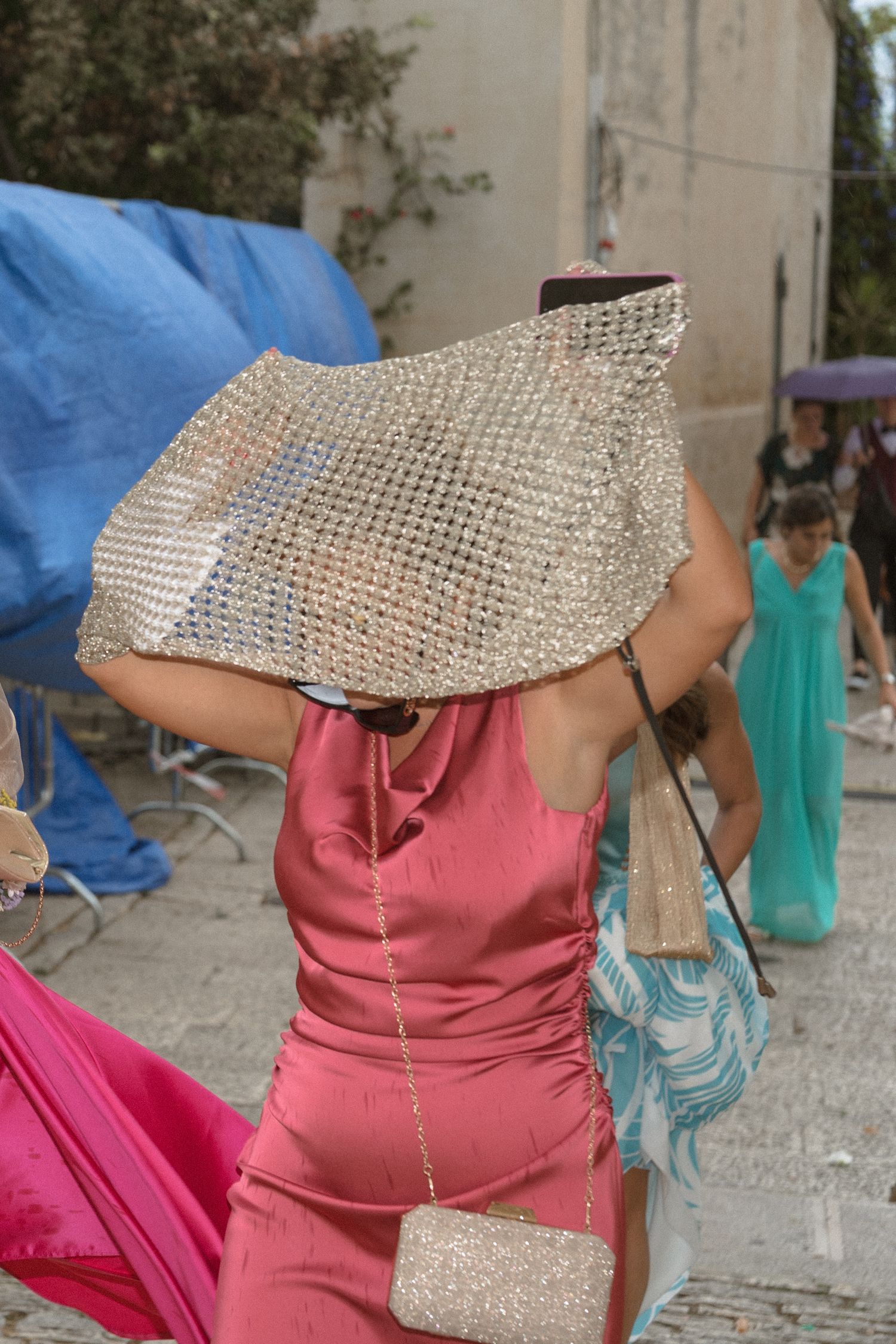 Woman in pink dress holds silver sequined clutch over her head outdoors at a formal event.