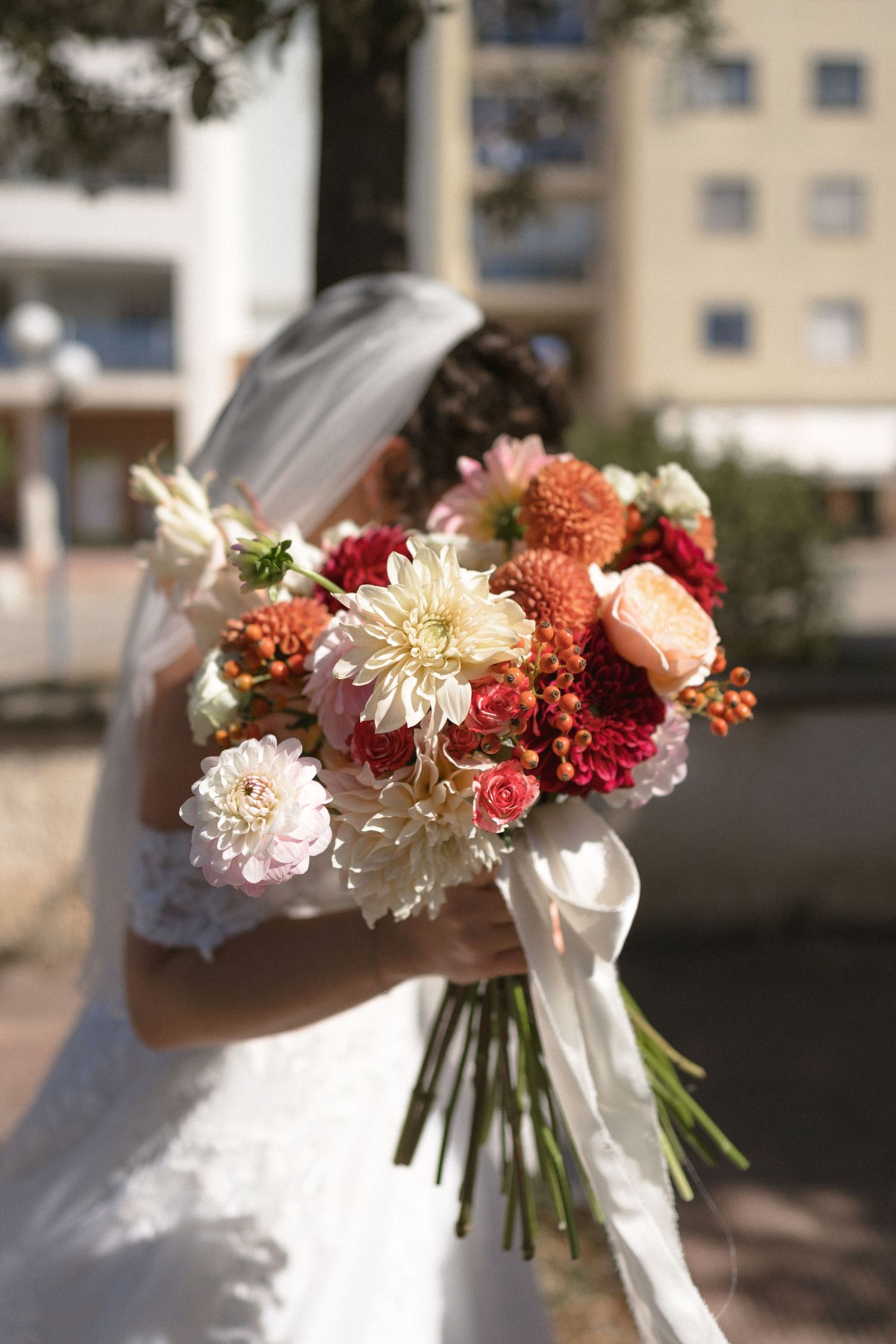 Bride in white veil holds vibrant bouquet of dahlias, roses, and orange flowers outdoors.