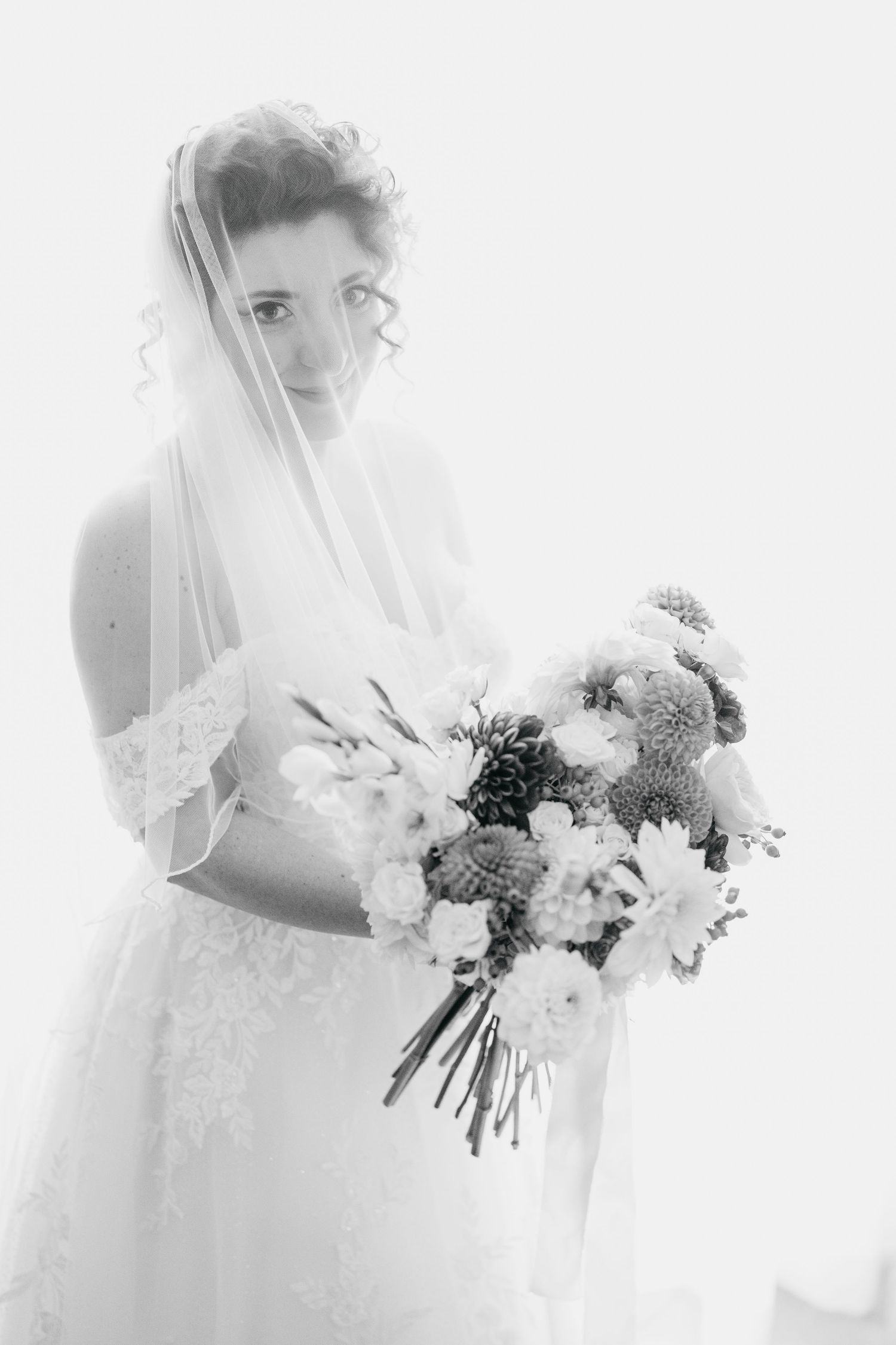 Elegant bride in lace gown and veil holding a bouquet in a soft black and white portrait.