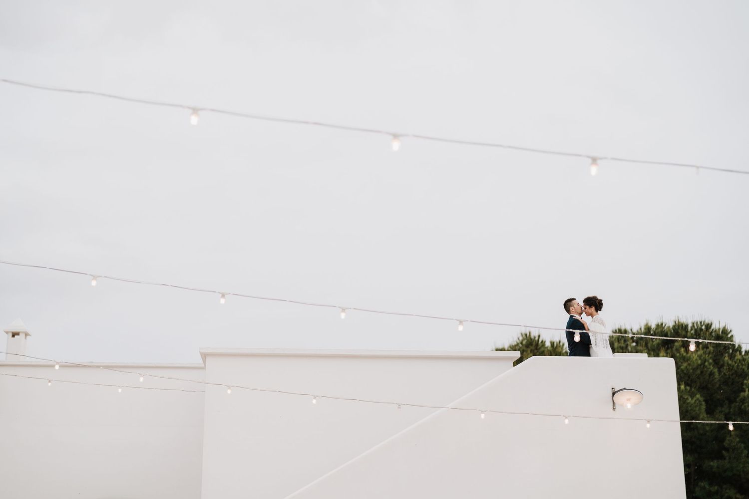 A distant romantic silhouette of a couple standing on a white rooftop against a bright sky.
