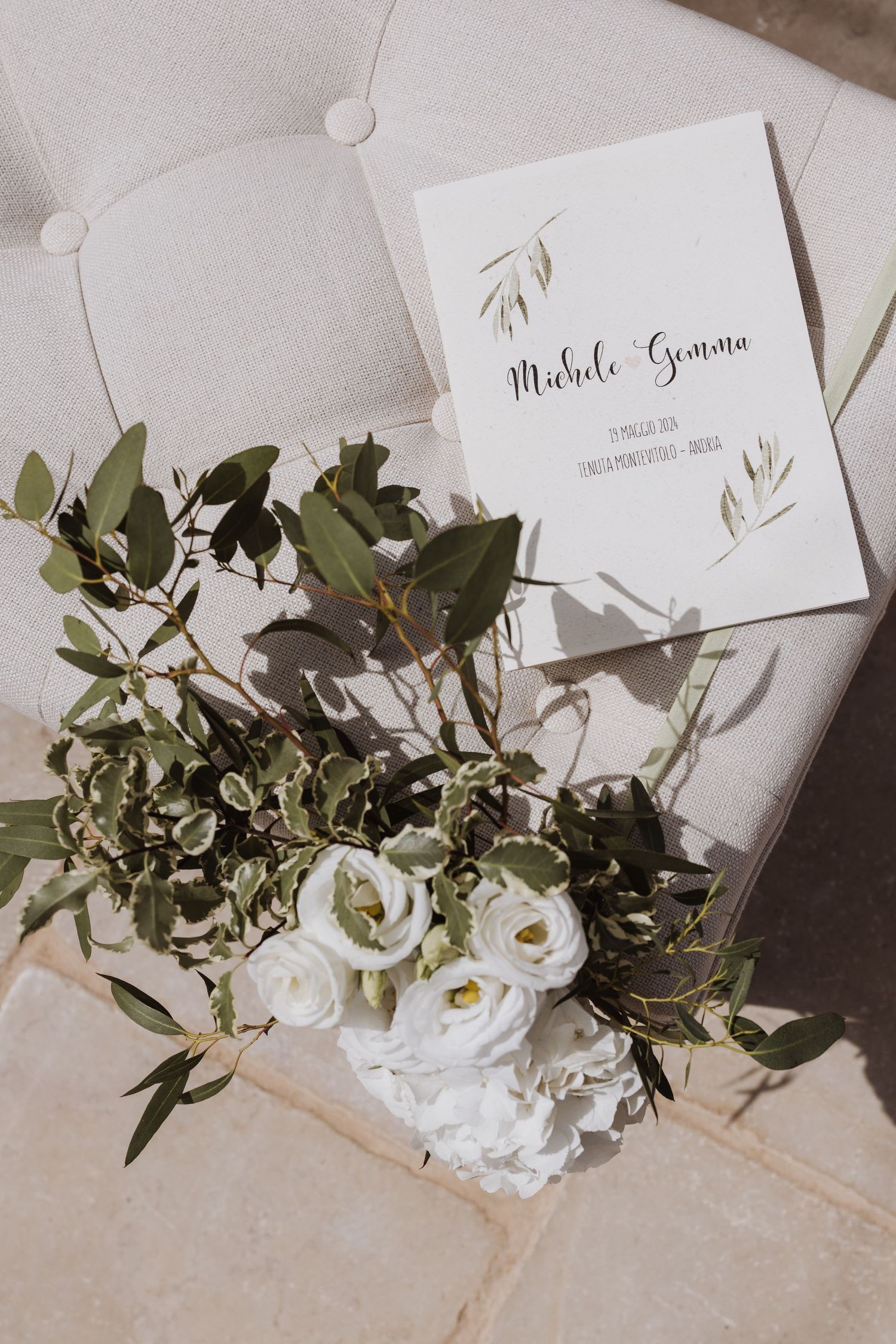 White floral bouquet with greenery and wedding invitation on a tufted light gray surface.