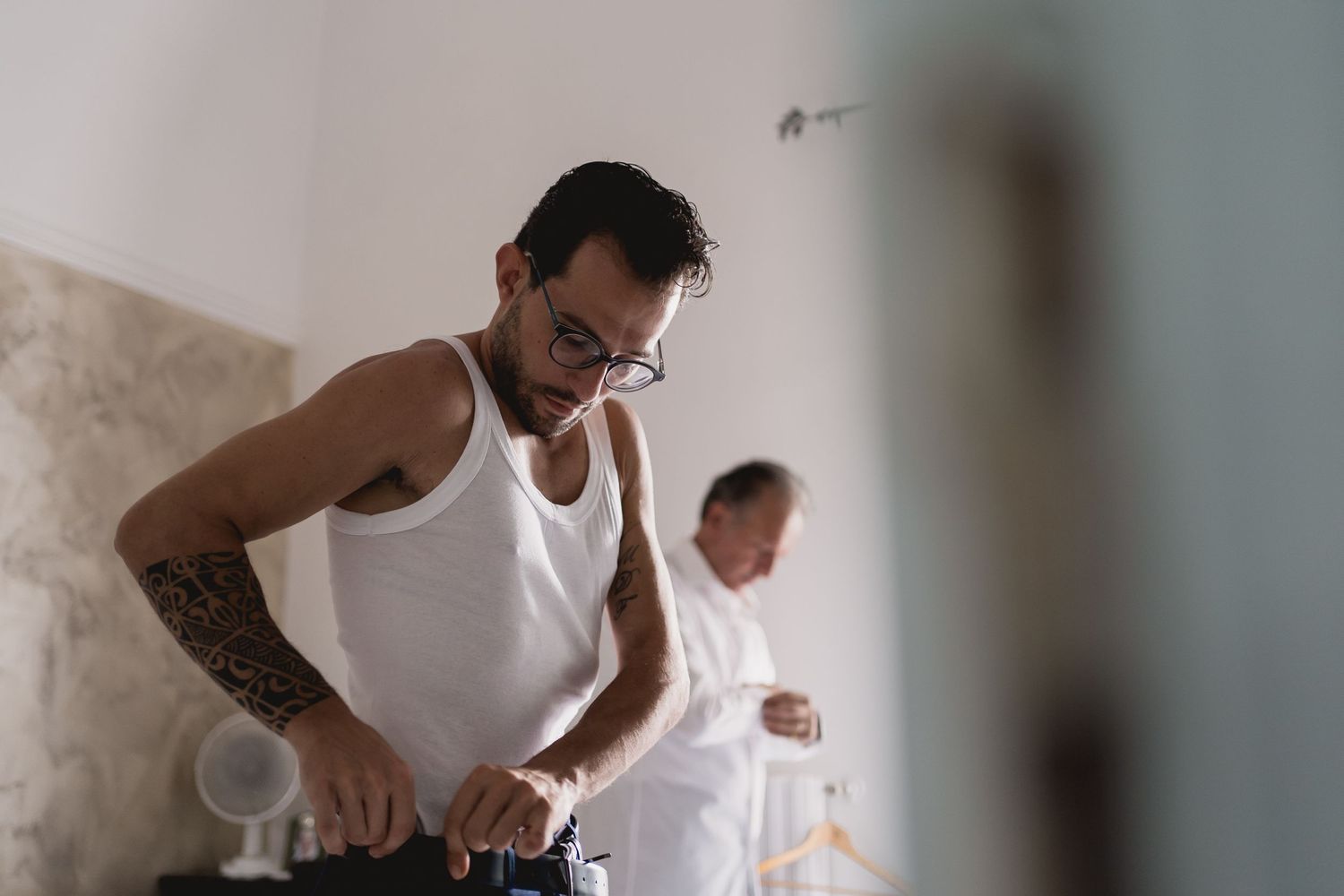 Man in white tank top getting ready in sunlit room.