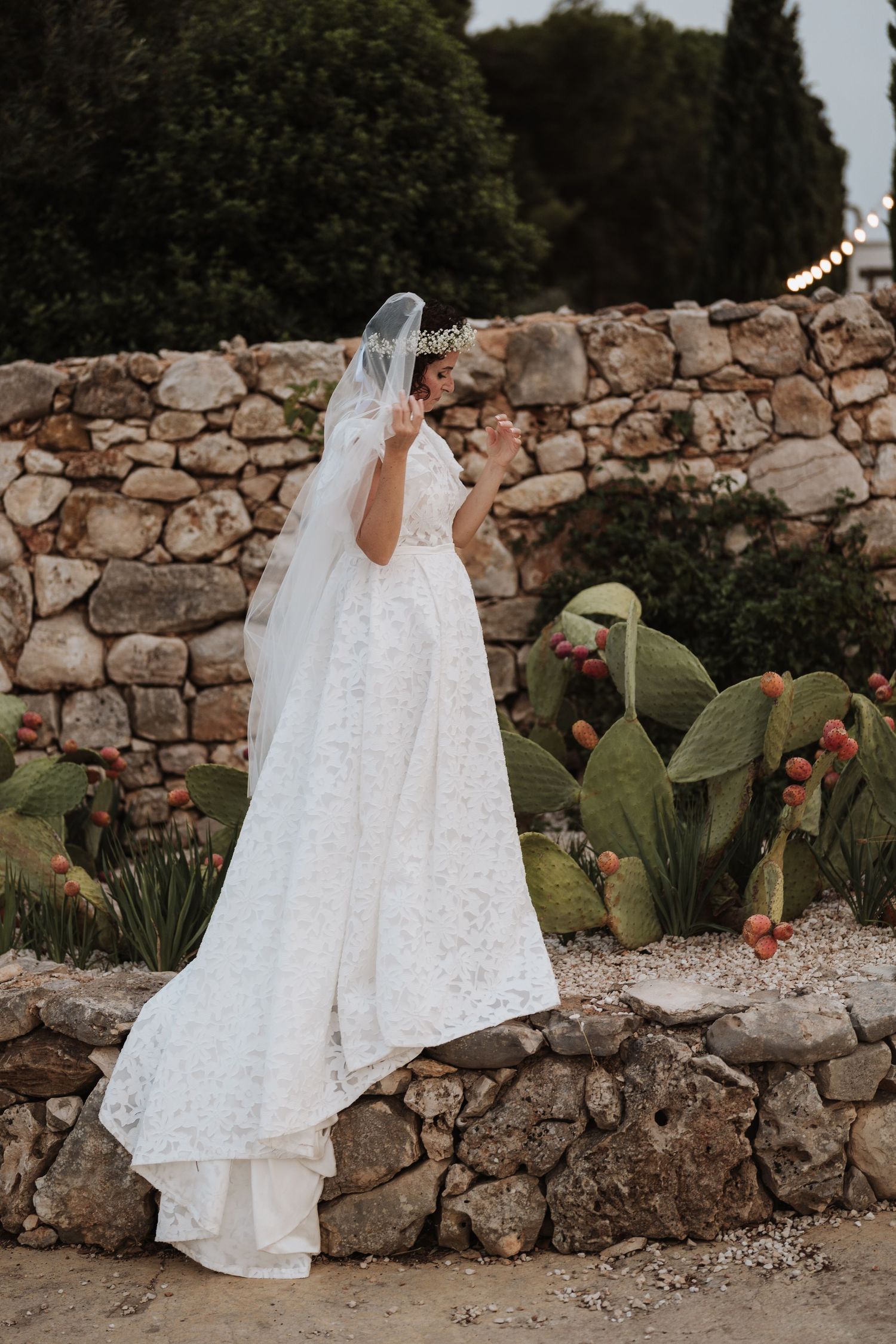 An elegant white lace wedding dress and veil draped over a rustic stone wall with flowering cacti in the background.
