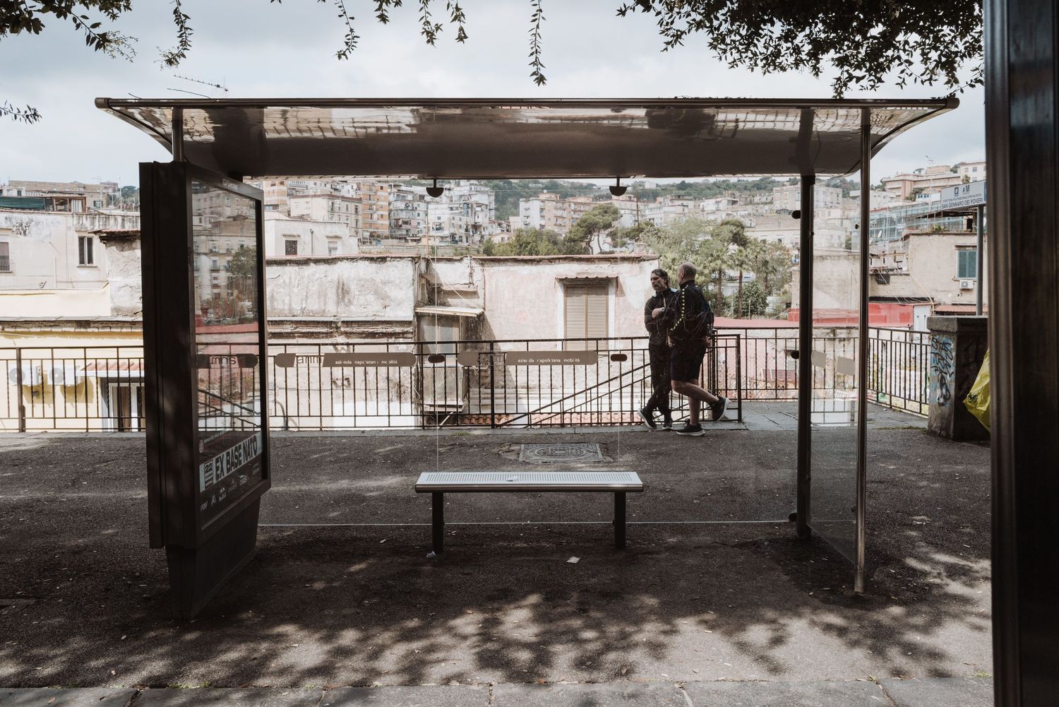 A person stands under a covered urban bus shelter with a bench on a city terrace overlooking buildings and rooftops.